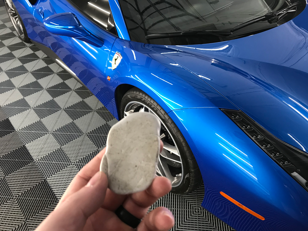 A person is holding a piece of clay in front of a blue car.