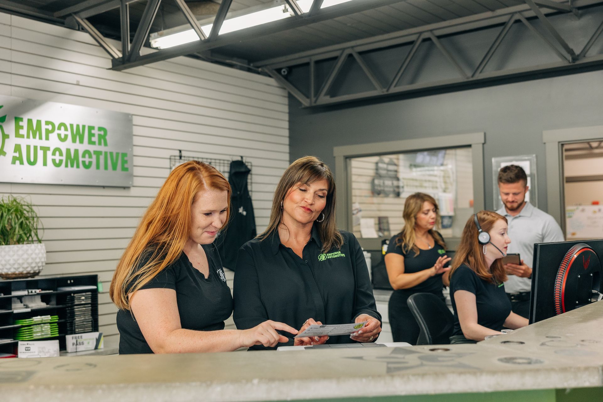 Inside an auto shop, women and a man staff a reception desk, discussing paperwork. | Empower Automotive