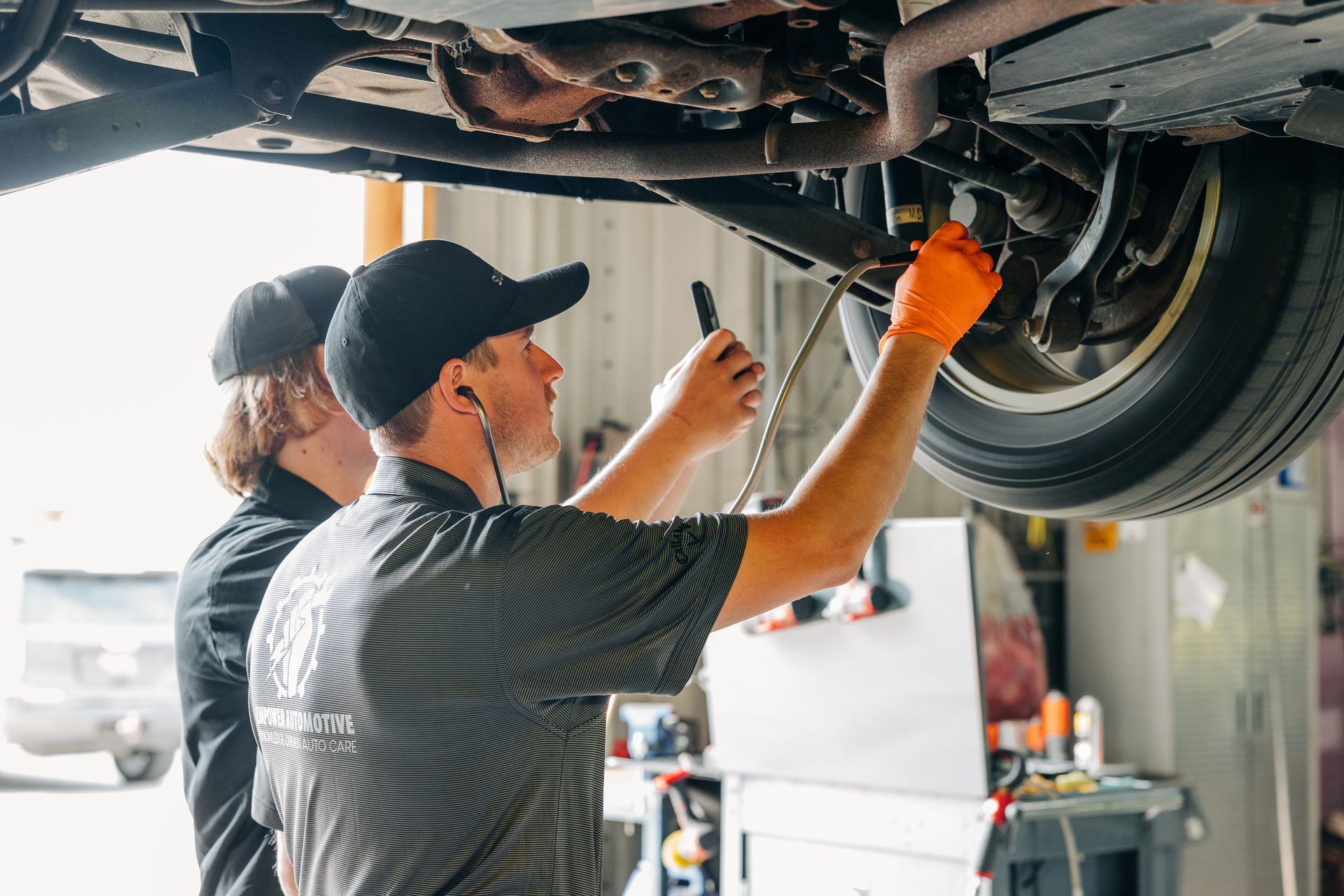 Two mechanics inspecting a car from below, one using a tool, wearing gloves and a cap. Garage setting. | Empower Automotive