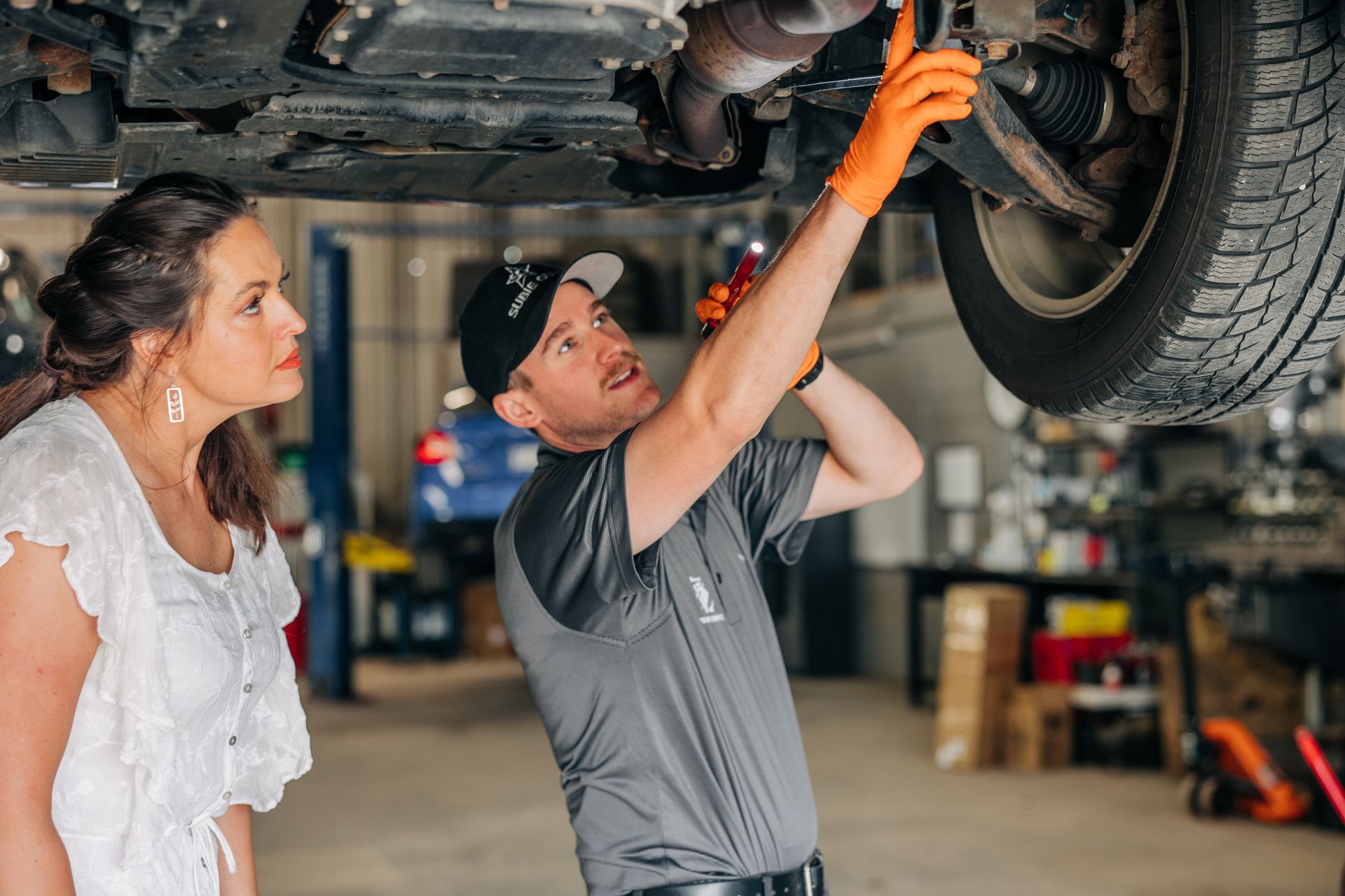Mechanic in orange gloves points at car undercarriage while explaining to a woman in a white top. | Empower Automotive