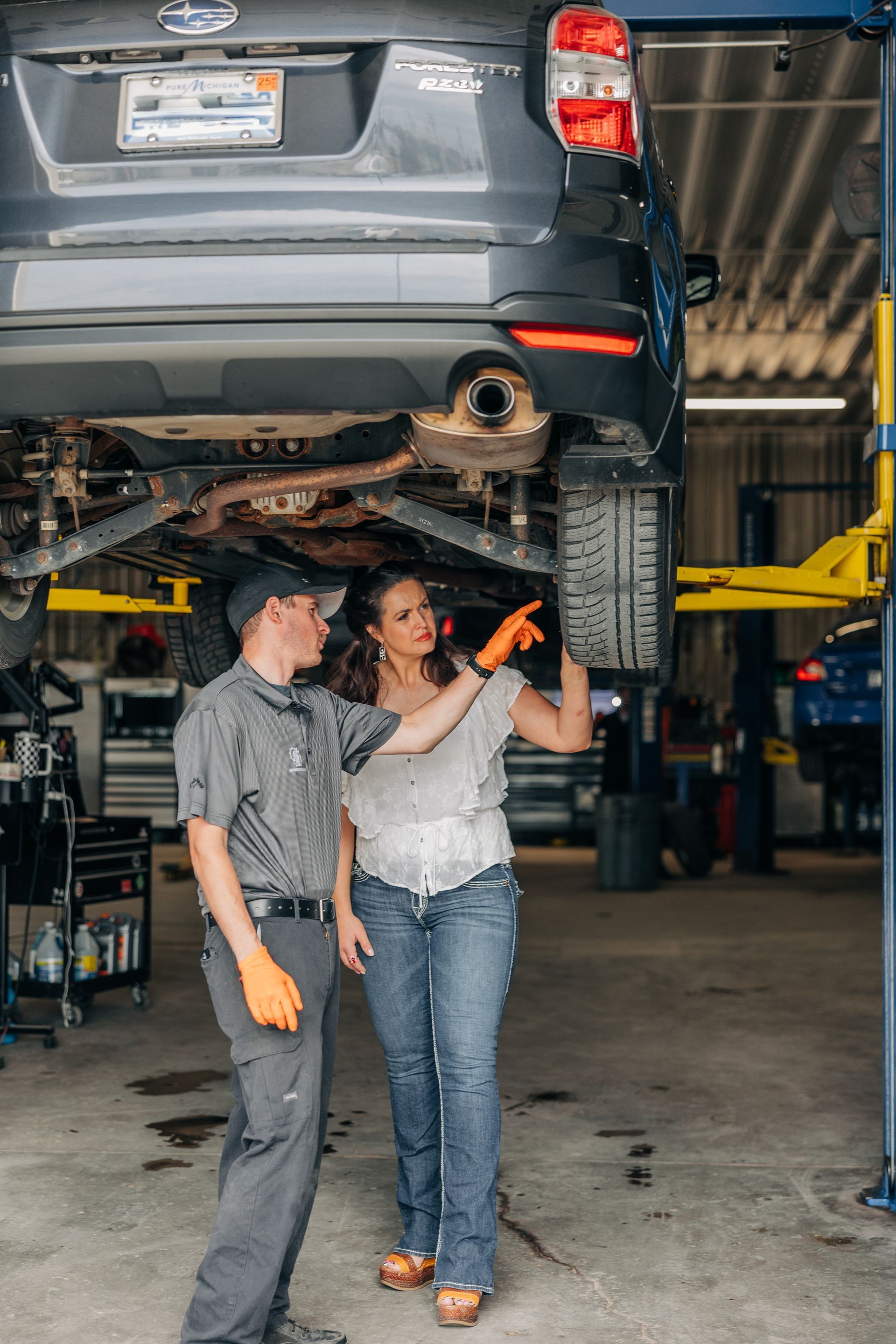 A mechanic and a woman discuss a car's undercarriage in a repair shop. The woman gestures at a component. | Empower Automotive