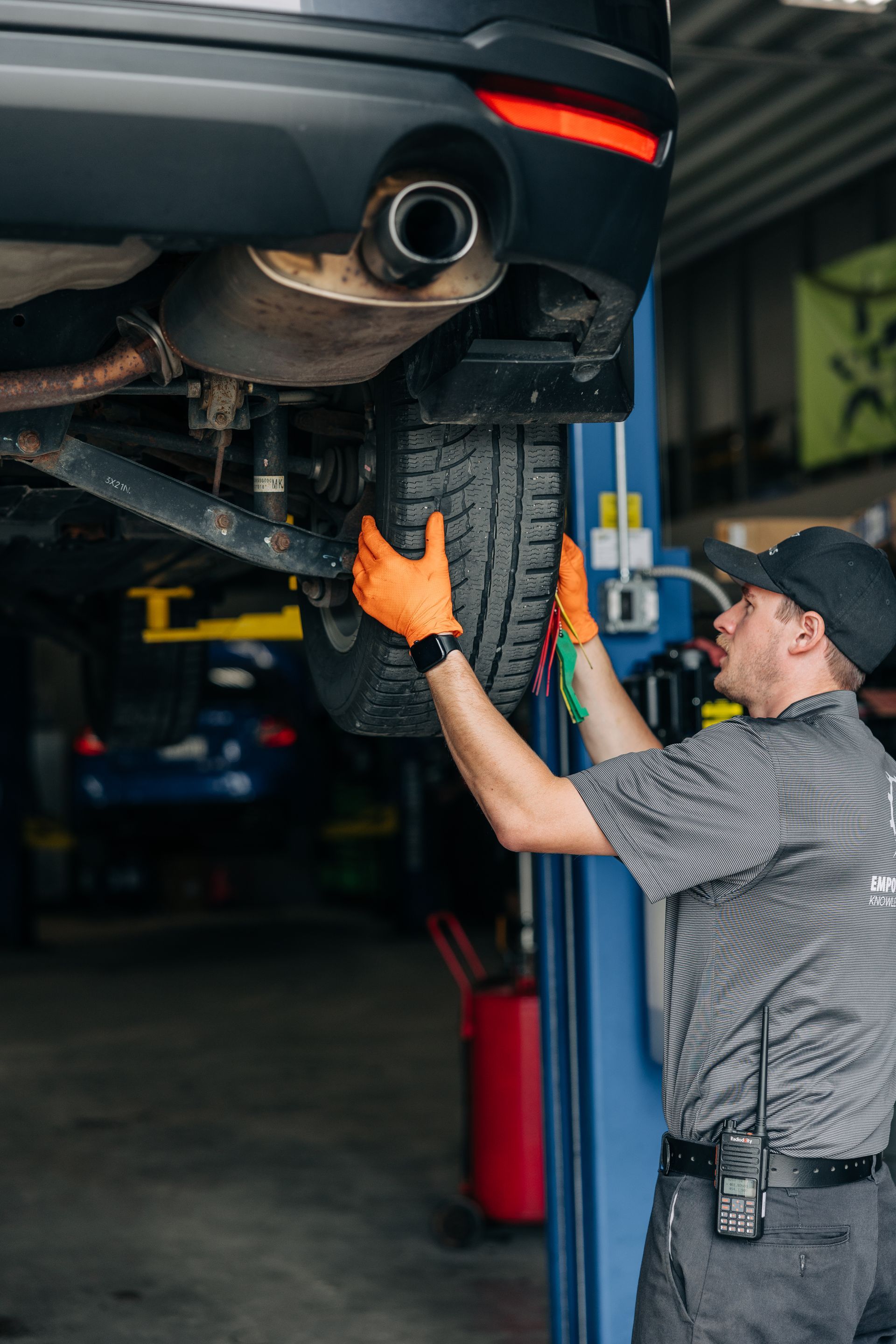 Mechanic inspecting car tire on lift, wearing orange gloves, in a garage. | Empower Automotive