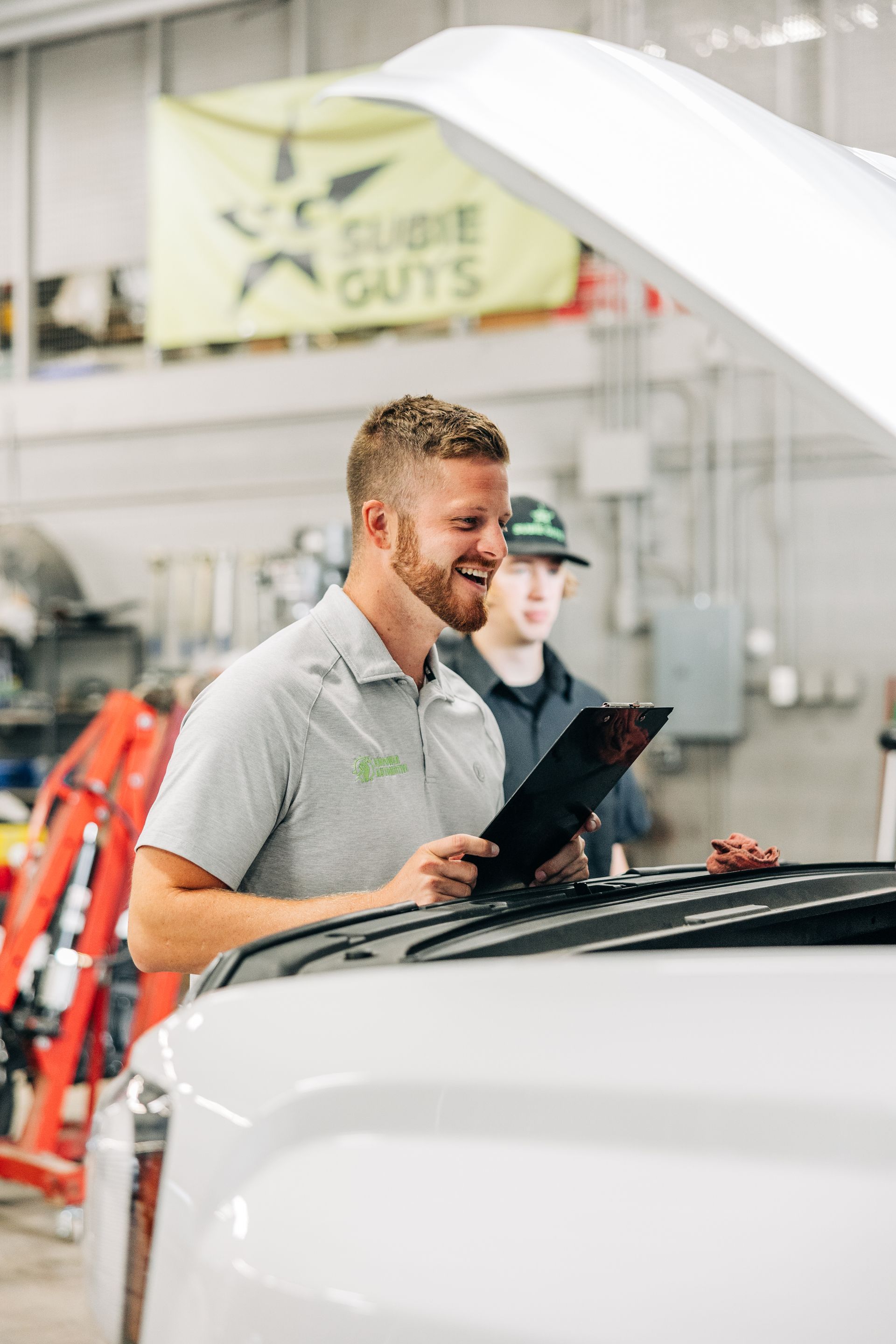 Mechanic smiling, reviewing paperwork, with car hood open, in a garage. Another person stands behind him.