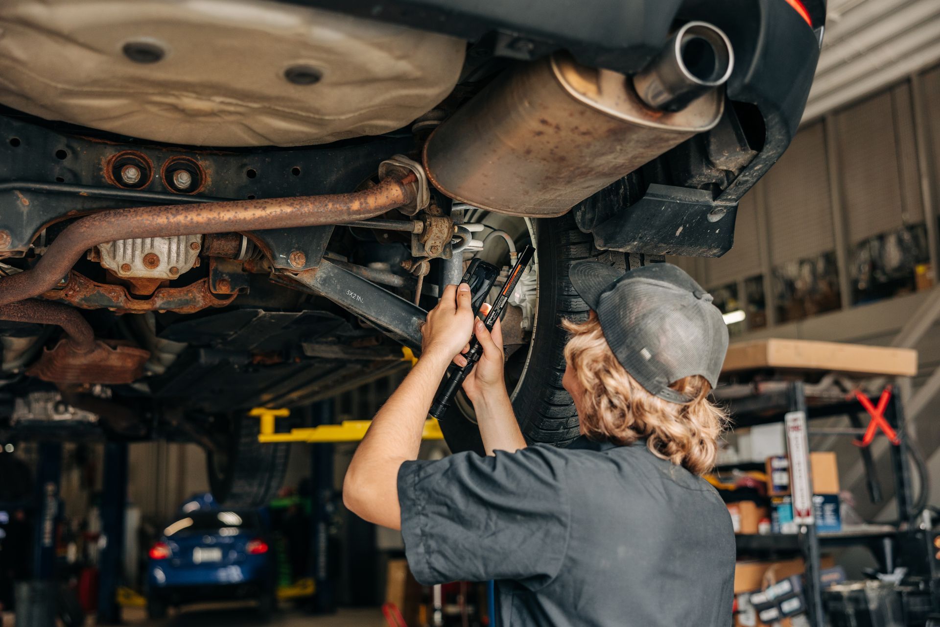 Mechanic working under a car on a lift, wearing a hat, with exhaust and suspension visible. | Empower Automotive