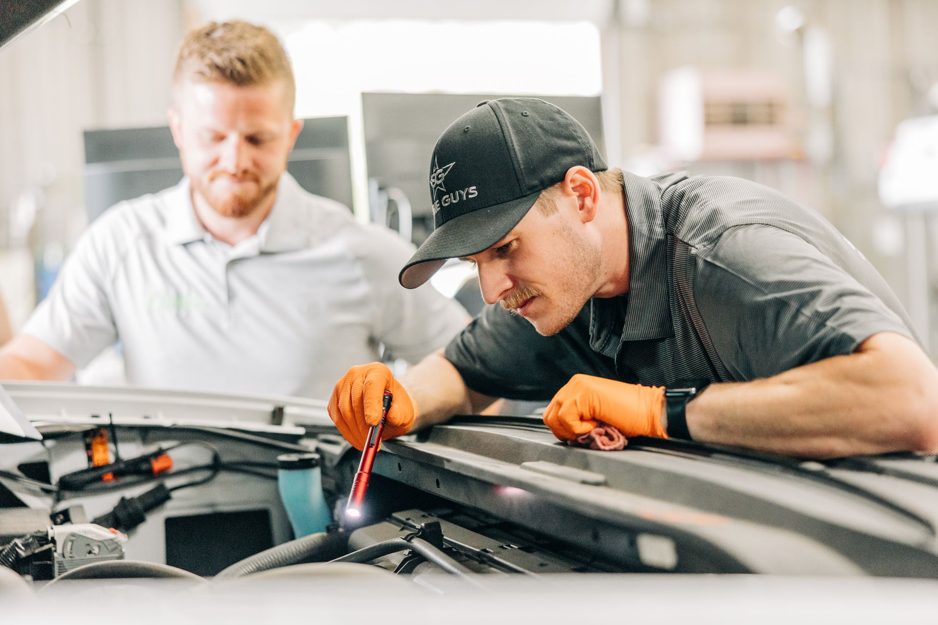 Two mechanics inspecting a car engine. One uses a tool with orange gloves on, the other watches.