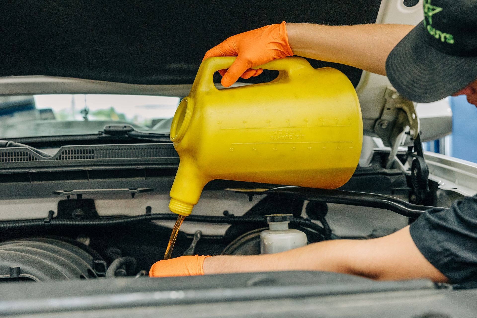 Mechanic pouring oil from a yellow jug into a car engine; wearing orange gloves.