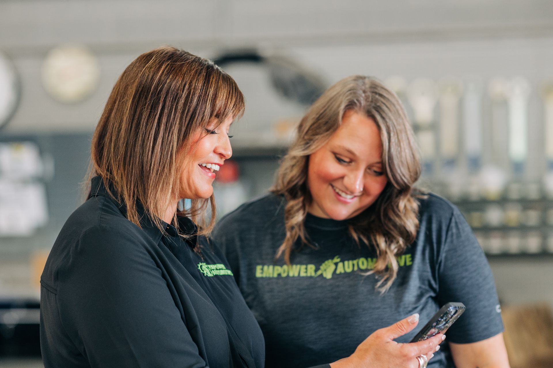 Two women looking at a phone, smiling. One wears a black shirt, the other a gray 