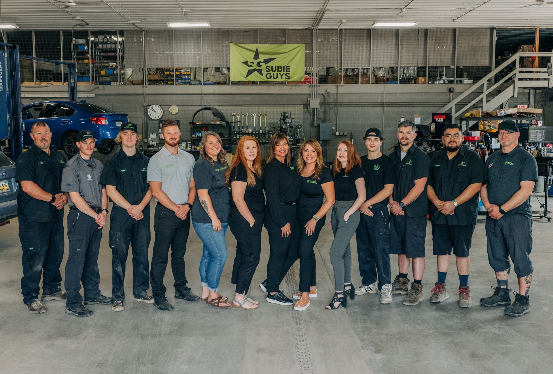 Group of mechanics in a garage posing for a photo. They are smiling, wearing matching shirts, in front of their shop.
