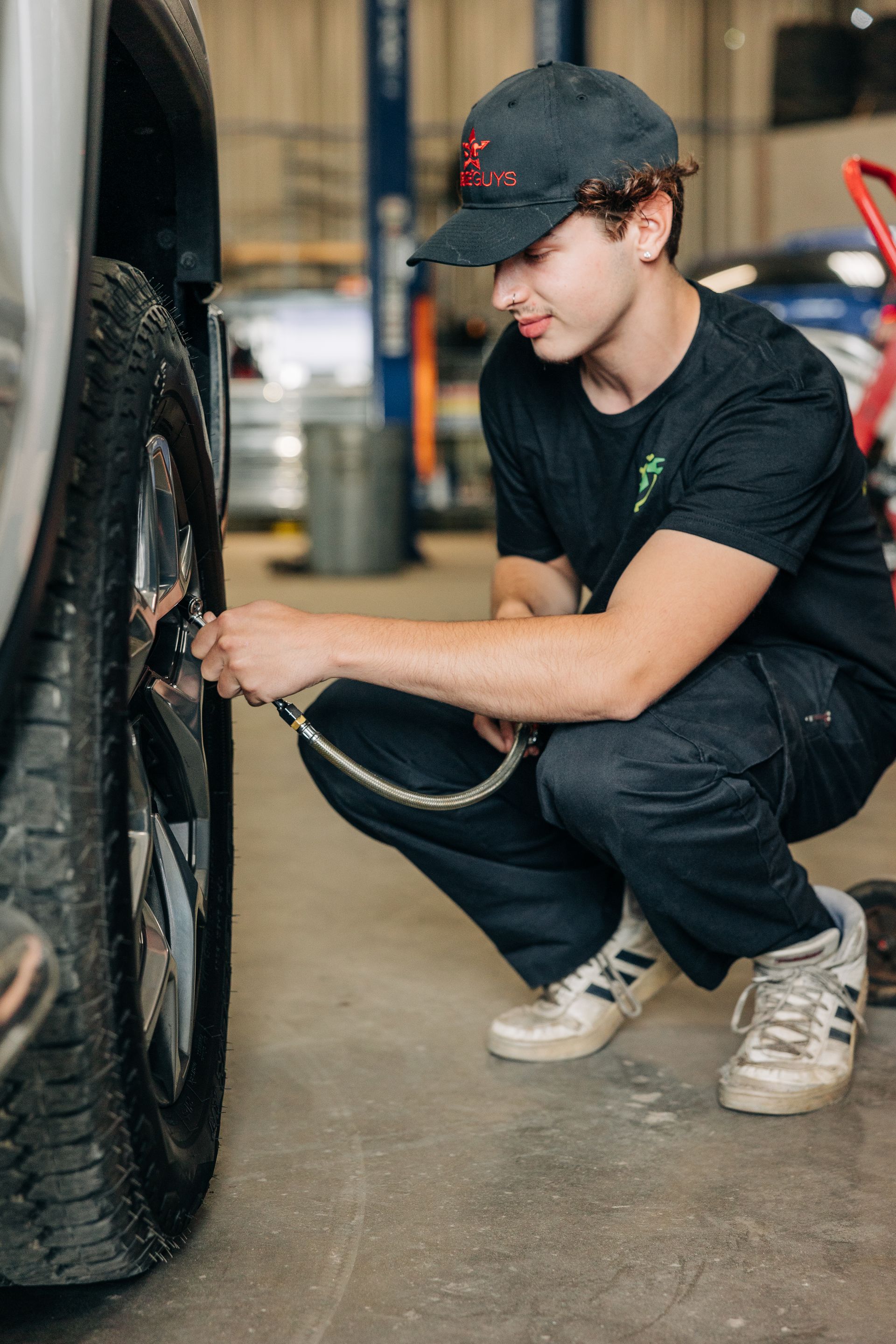 Man in cap inflates a tire in a garage. He is kneeling, wearing a dark t-shirt and pants, and white sneakers.