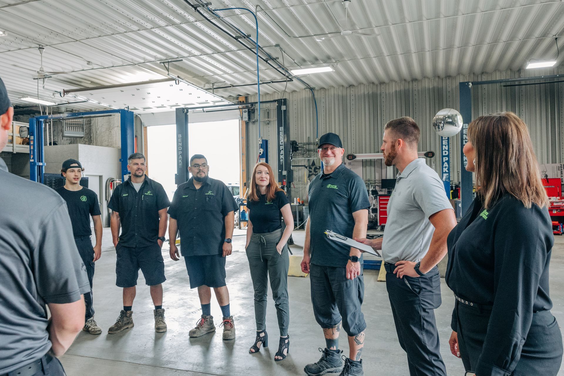Group of people in a garage, likely for a meeting. Several wearing matching green shirts, some gesturing and smiling. | Empower Automotive