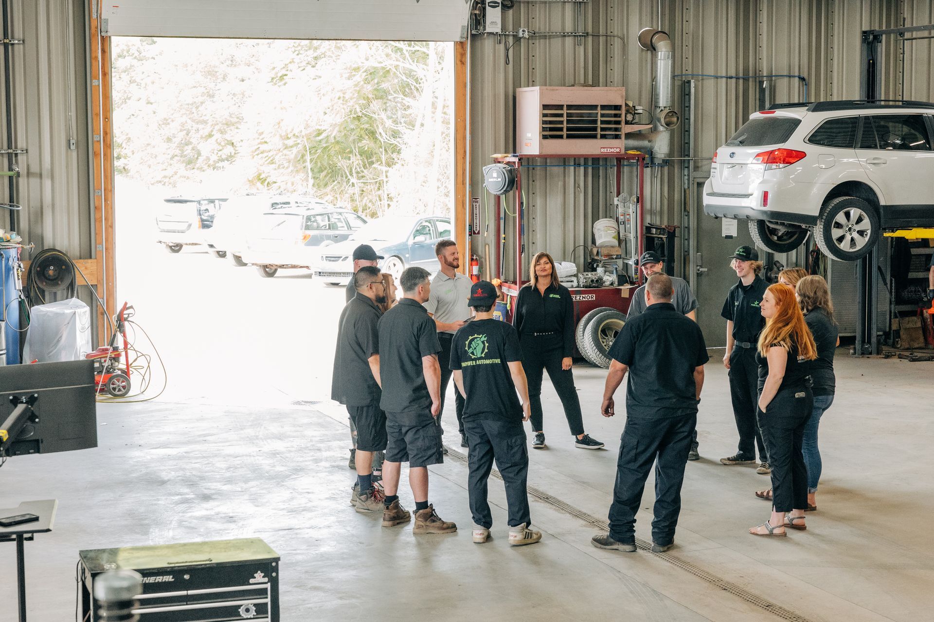 Group of people in a garage, gathered in a circle. Cars on lift, open door to outdoors. | Empower Automotive