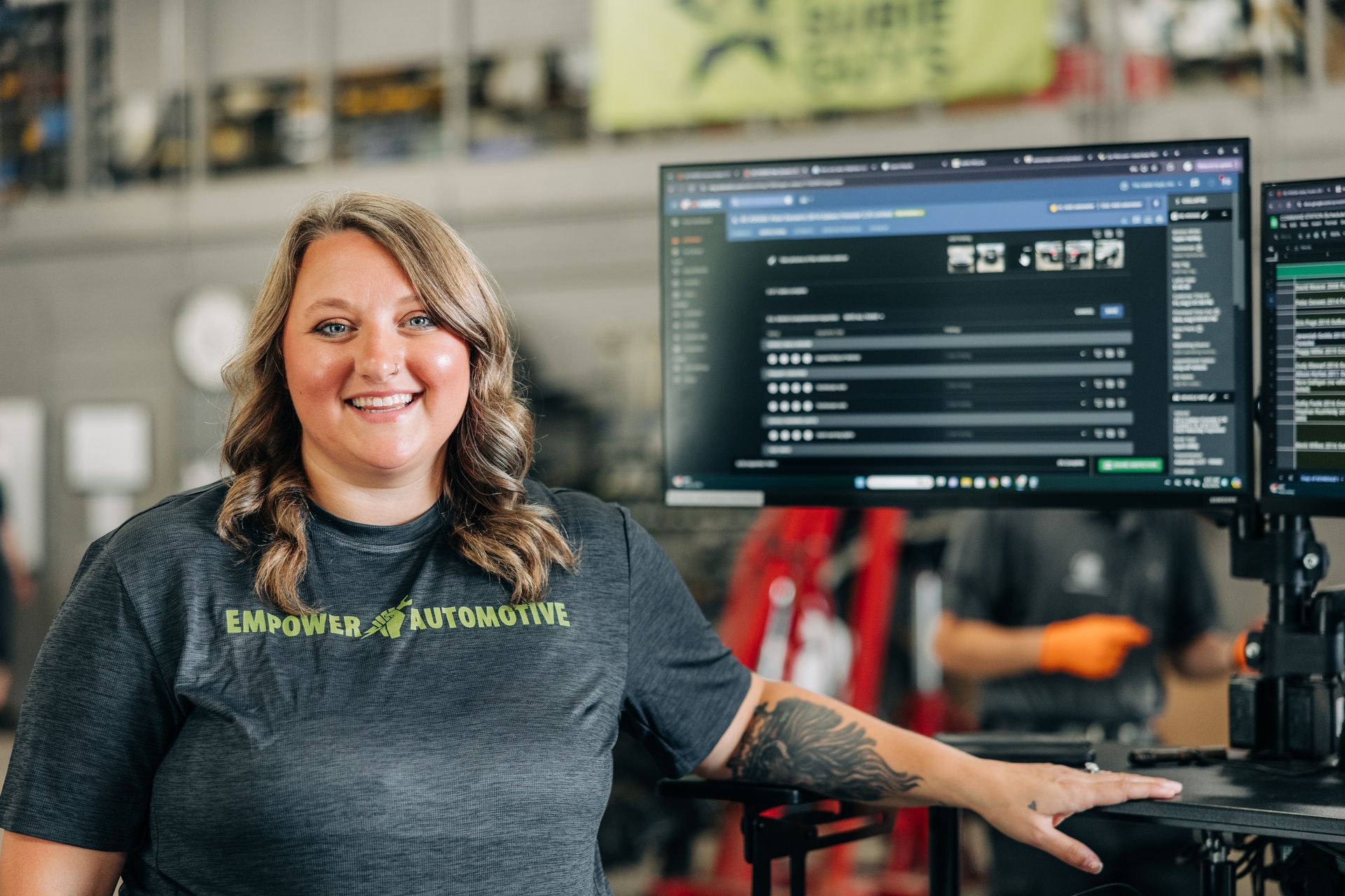 Woman with a tattoo smiles in front of a computer screen in an auto repair shop.