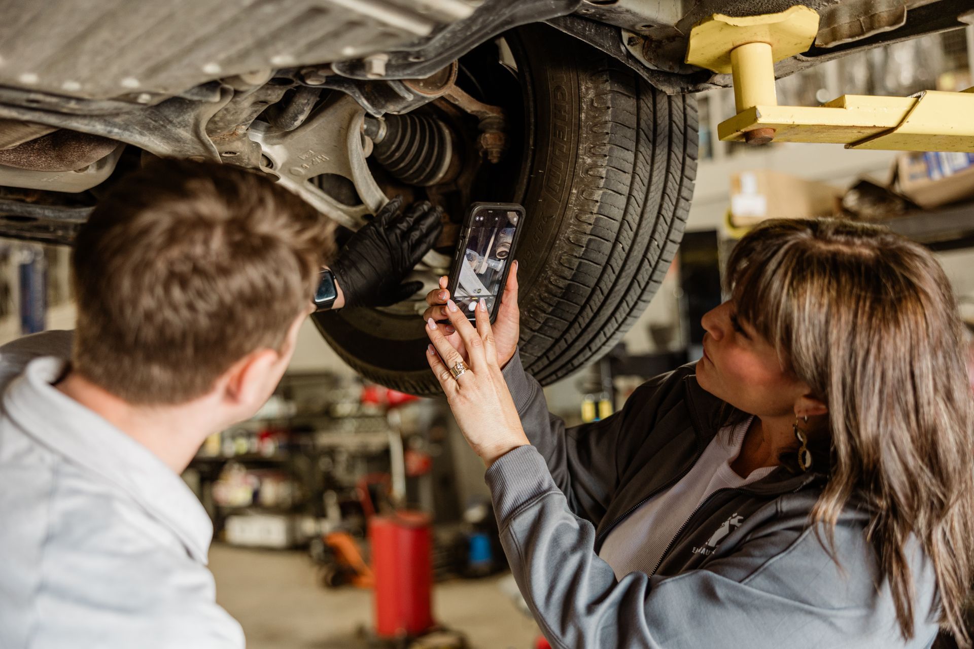 Woman takes photo of car undercarriage with phone, man looks on.