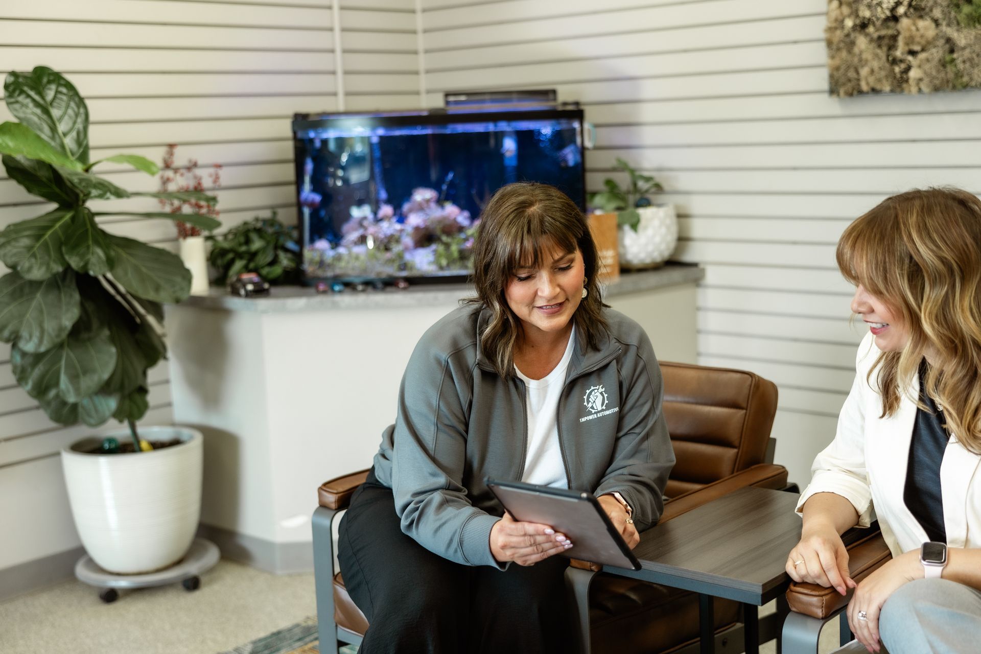 Two women in an office setting. One shows the other a tablet while sitting in chairs near an aquarium.
