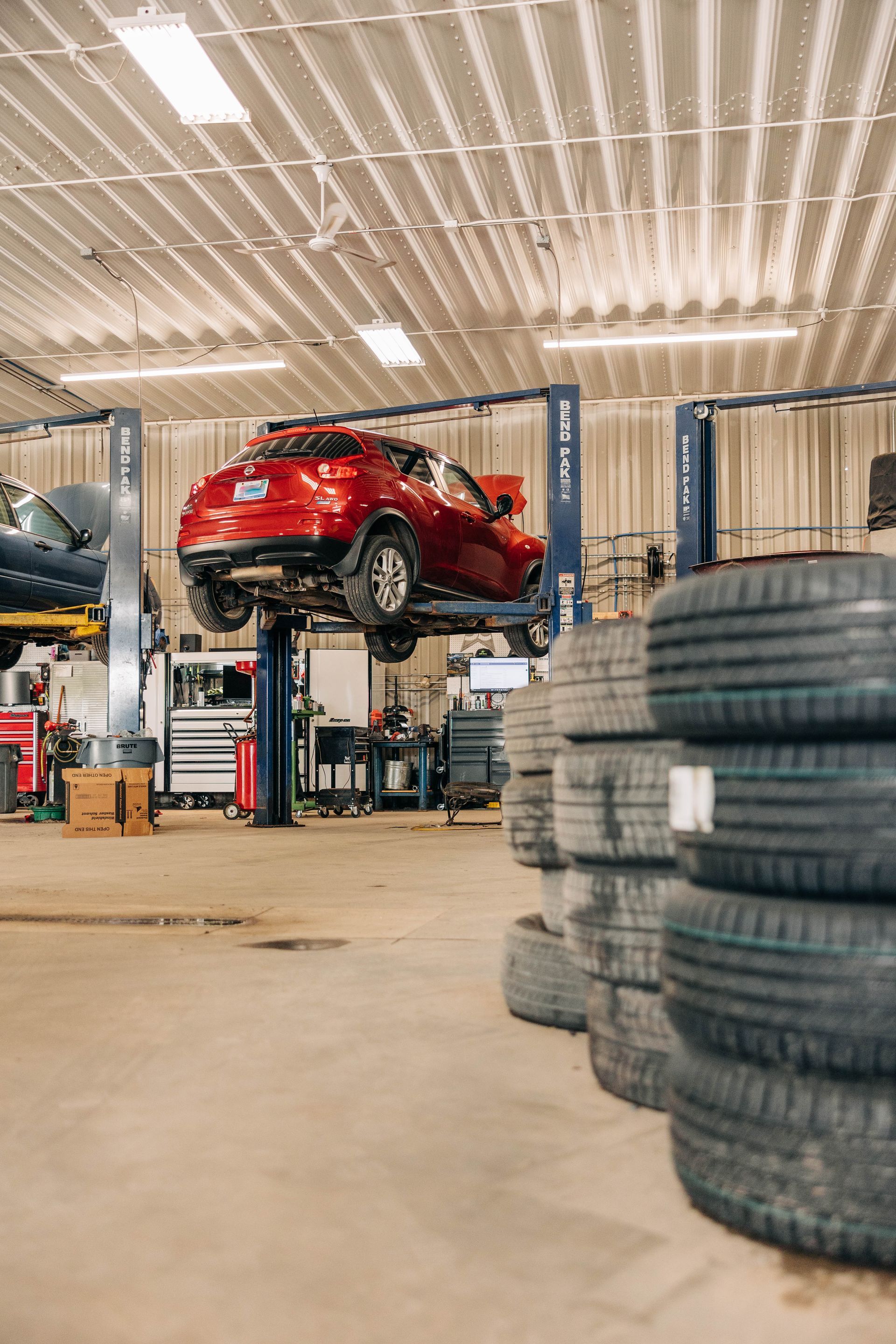 Red car elevated on a lift in a repair shop, with stacked tires in the foreground. | Empower Automotive