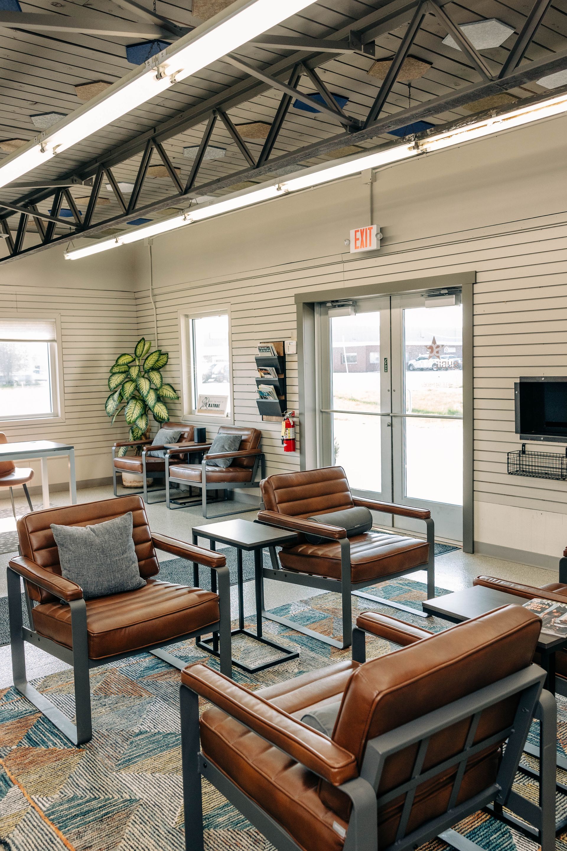 Cozy waiting area with brown leather chairs, small tables, rug, windows, and an exit door. | Empower Automotive