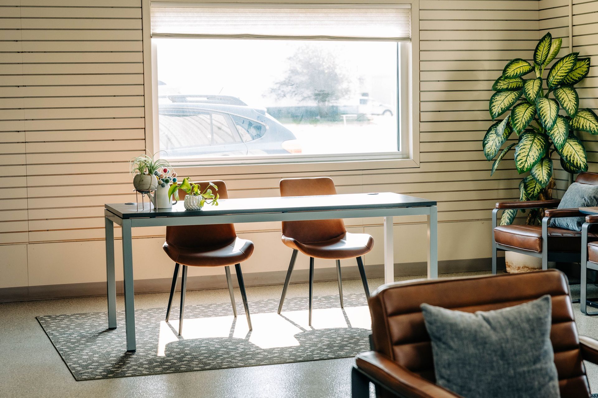 Desk with chairs, window, and potted plant in a well-lit room with brown leather furniture. | Empower Automotive