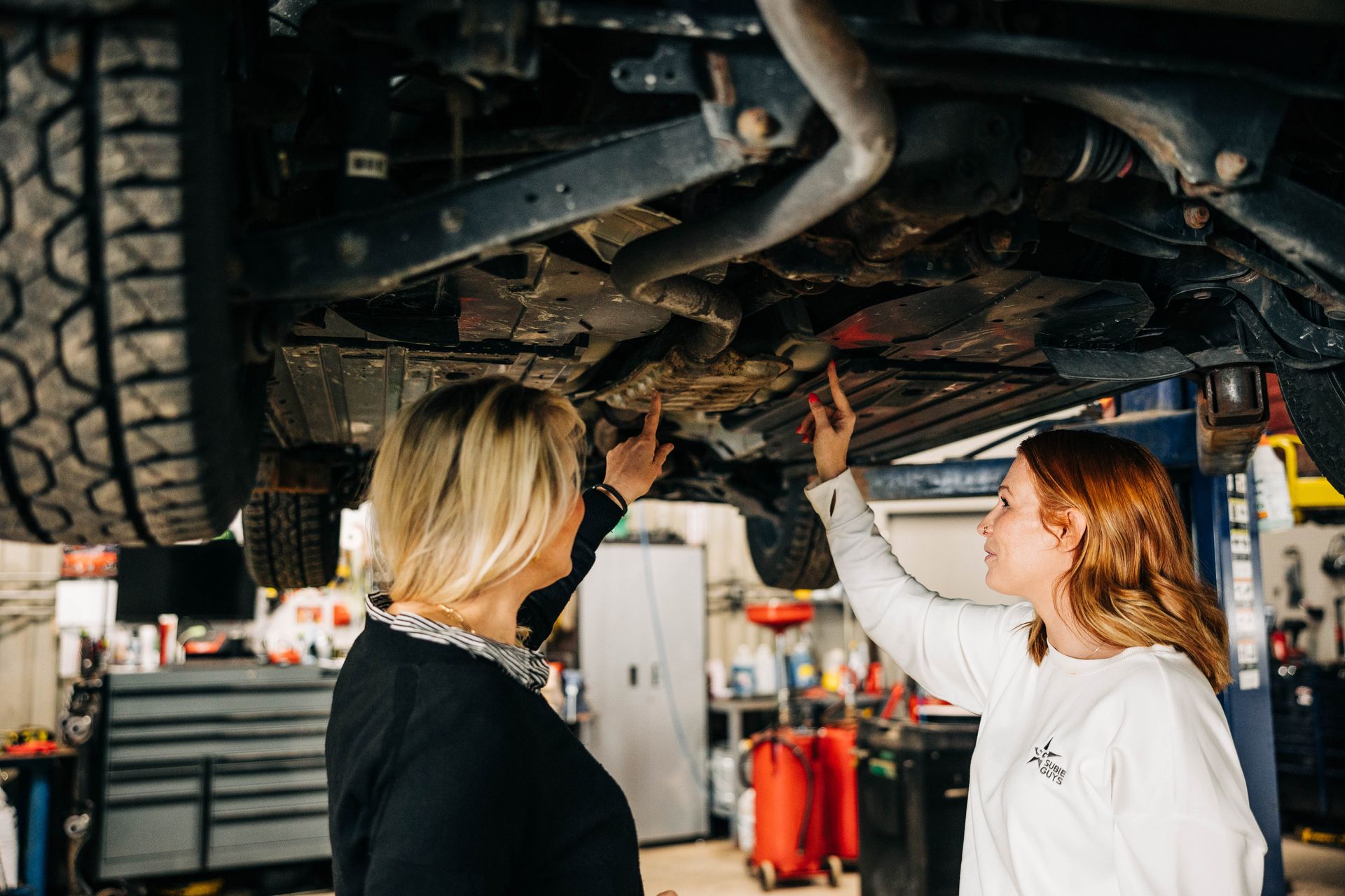 Two women pointing at the undercarriage of a vehicle in a garage. | Empower Automotive