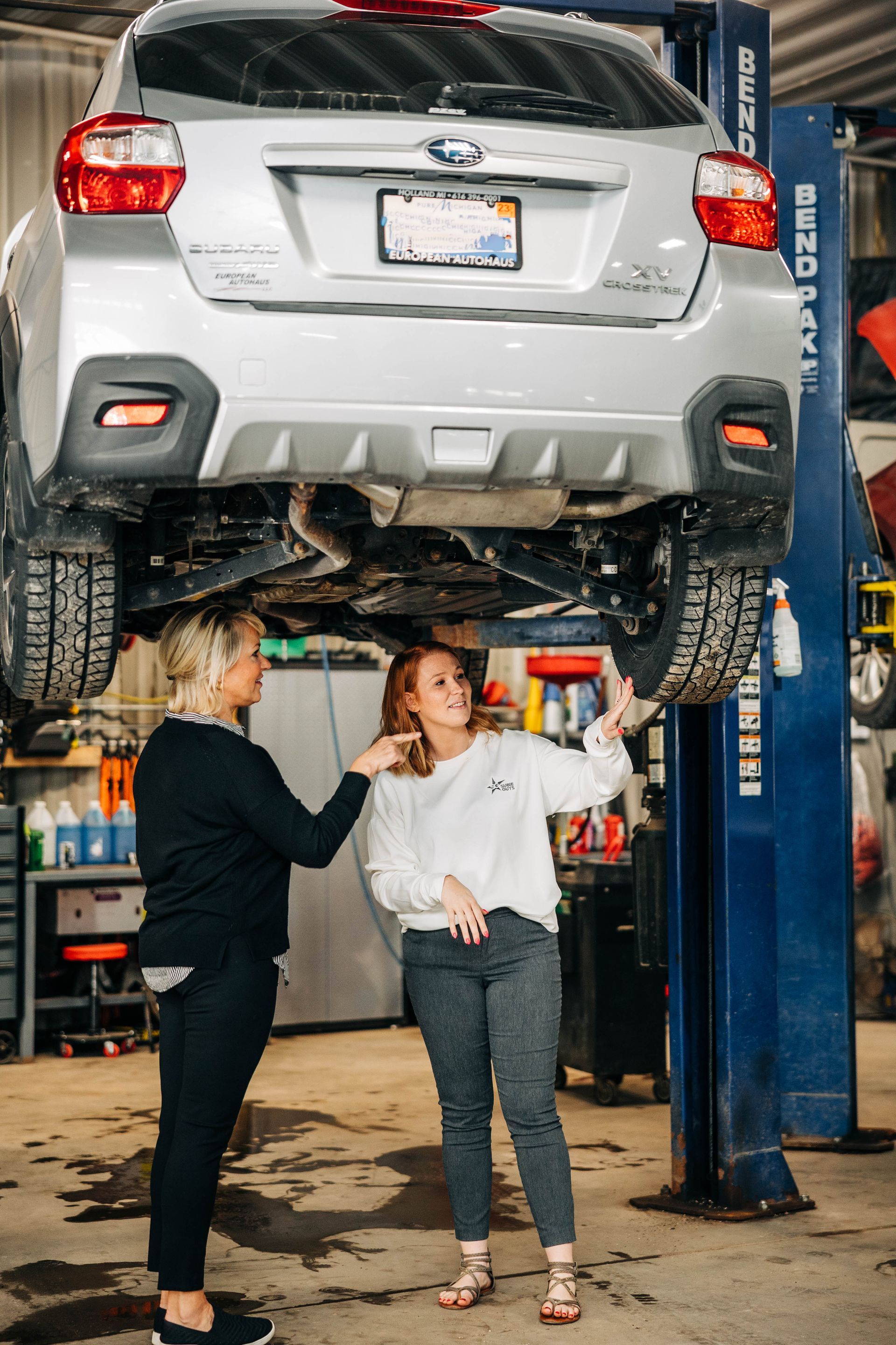 Two women examining the underside of a silver Subaru Crosstrek lifted on a blue car lift in a garage. | Empower Automotive
