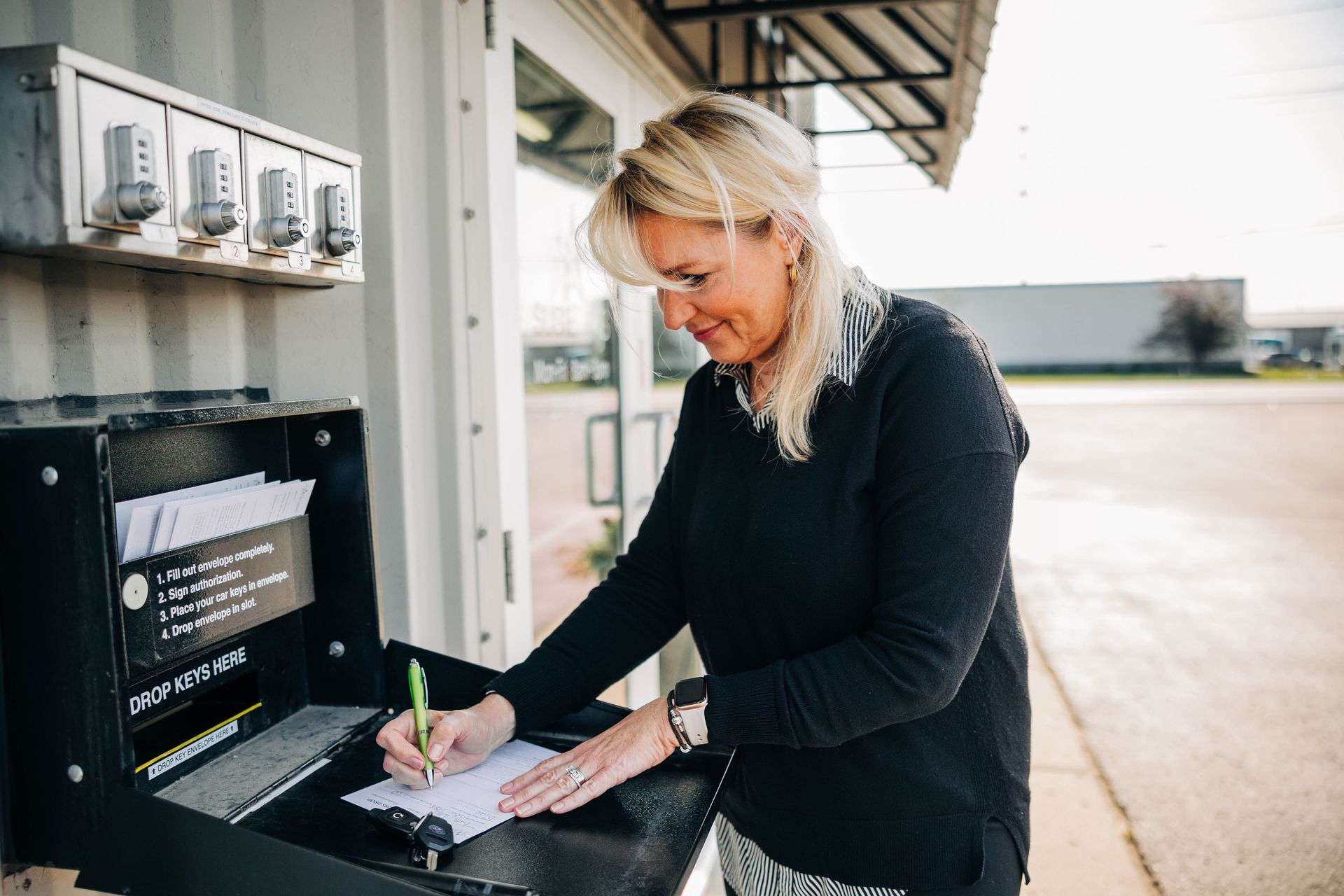 Woman writing at a black outdoor desk near gray metal lockers, outside. | Empower Automotive