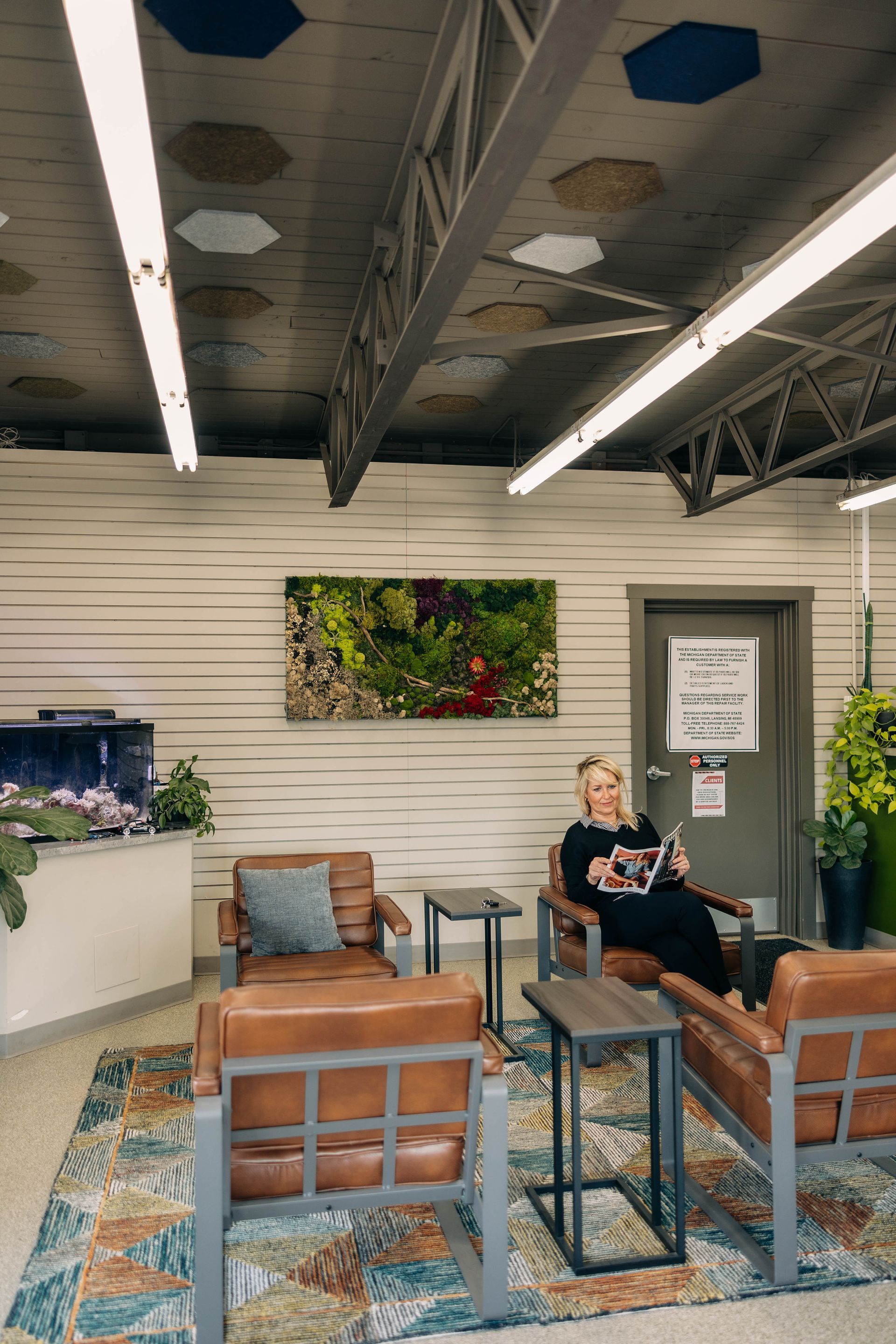 Woman in a waiting room reading magazine, brown leather chairs, large rug, greenery, and a fish tank. | Empower Automotive