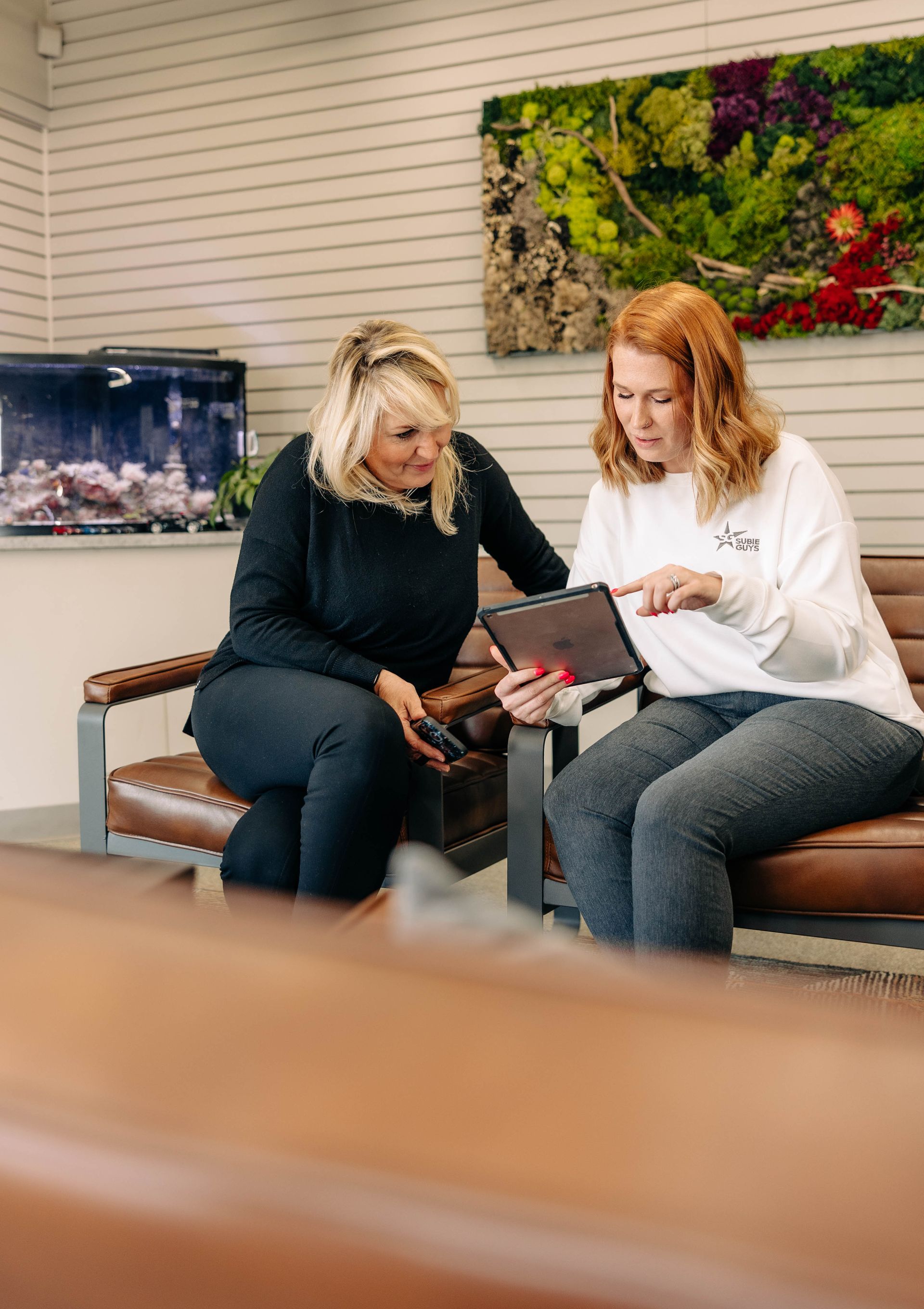 Two women looking at a tablet, sitting on a brown bench. One points at the screen, a waiting room setting. | Empower Automotive