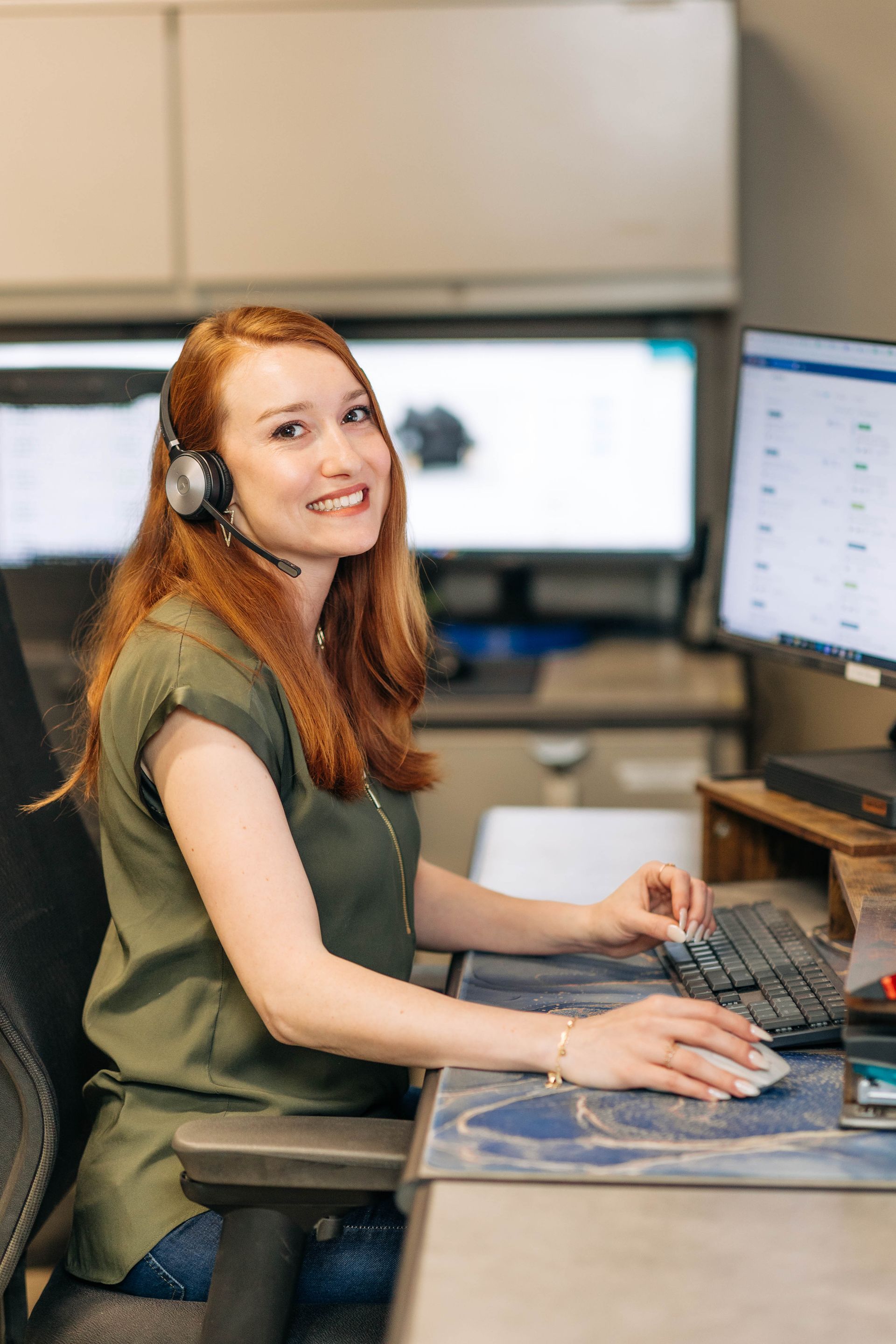 Woman with red hair wearing a headset, smiling, working at a computer in an office. | Empower Automotive