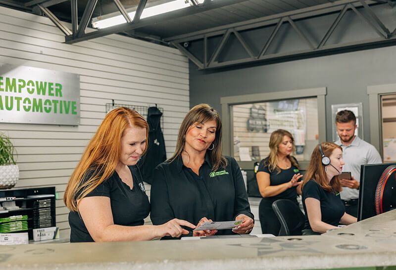 Employees at Empower Automotive in a garage setting; two women examining paperwork at the counter, others in the background.
