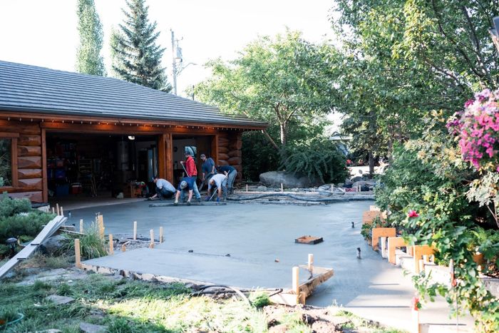 Construction workers pouring concrete for a driveway near a wooden building; trees and plants frame the scene.