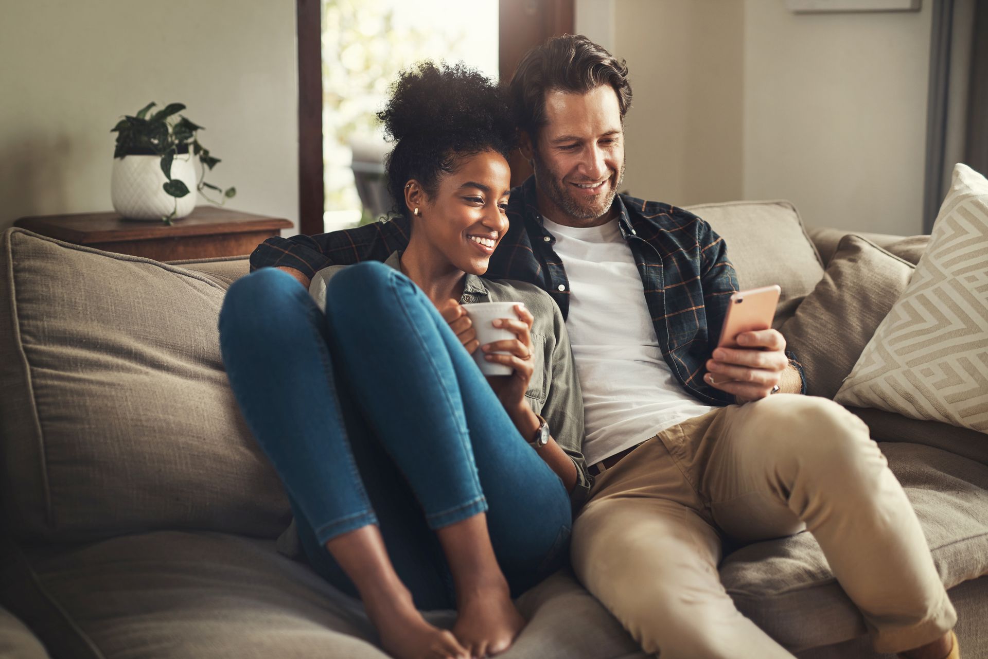 A smiling couple sits together on a couch, looking at a smartphone held by the man, while the woman holds a coffee mug.