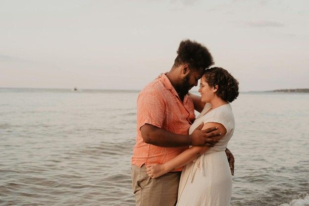 Couple embraces at the beach, heads touching. Ocean in background, overcast sky.