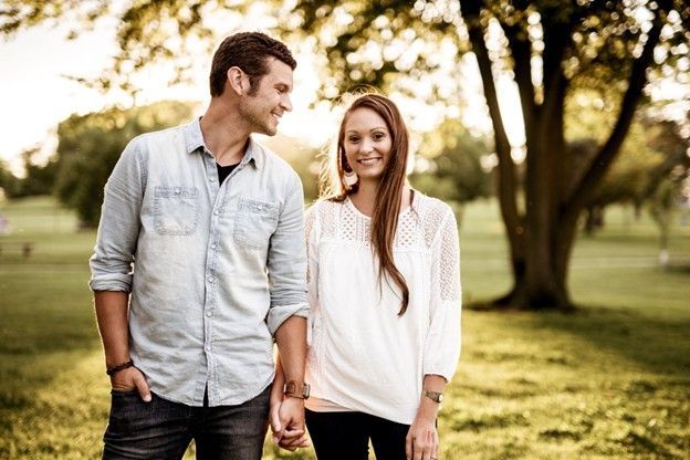 Couple holding hands, standing in a park, smiling. Man in denim shirt, woman in white top, soft sunlight.