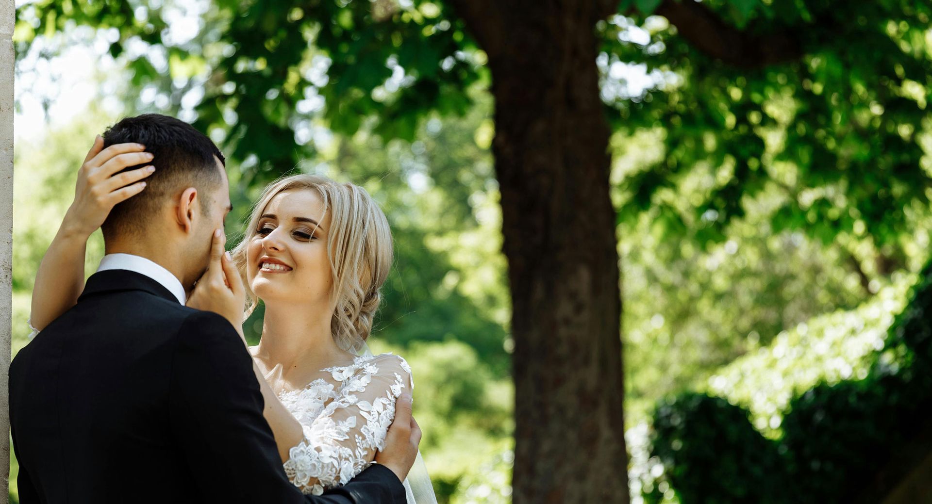 Bride and groom embrace outdoors, smiling. She wears a white lace dress; he wears a black suit.