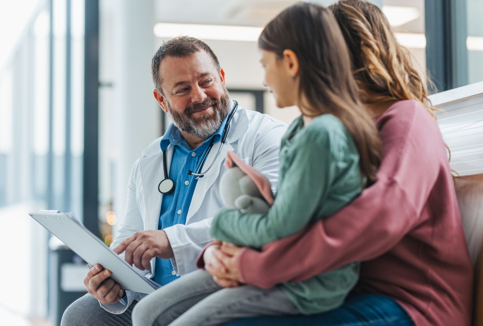 Doctor in white coat talks with woman and child in bright, modern medical office.