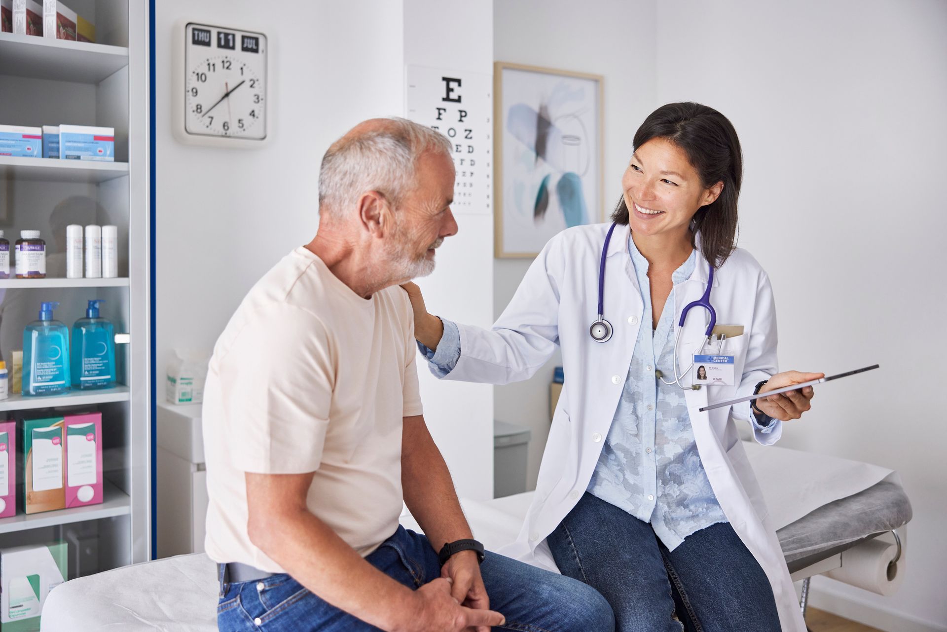 Doctor in white coat consults older patient in clinic with eye chart and medical supplies.