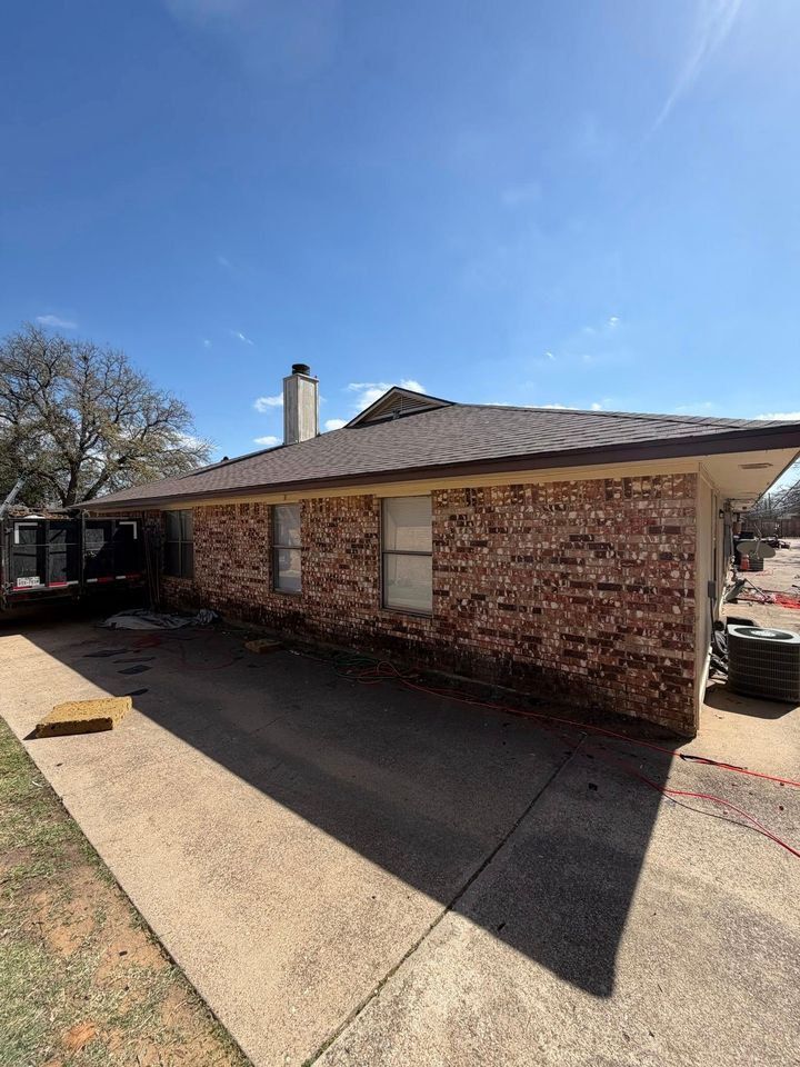 Brick house exterior with shingled roof, concrete patio, and clear blue sky.