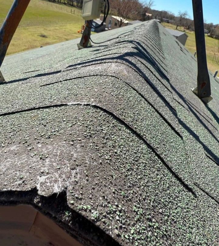 Damaged shingled roof with a long ridge hump and visible warped sections on a sunny day
