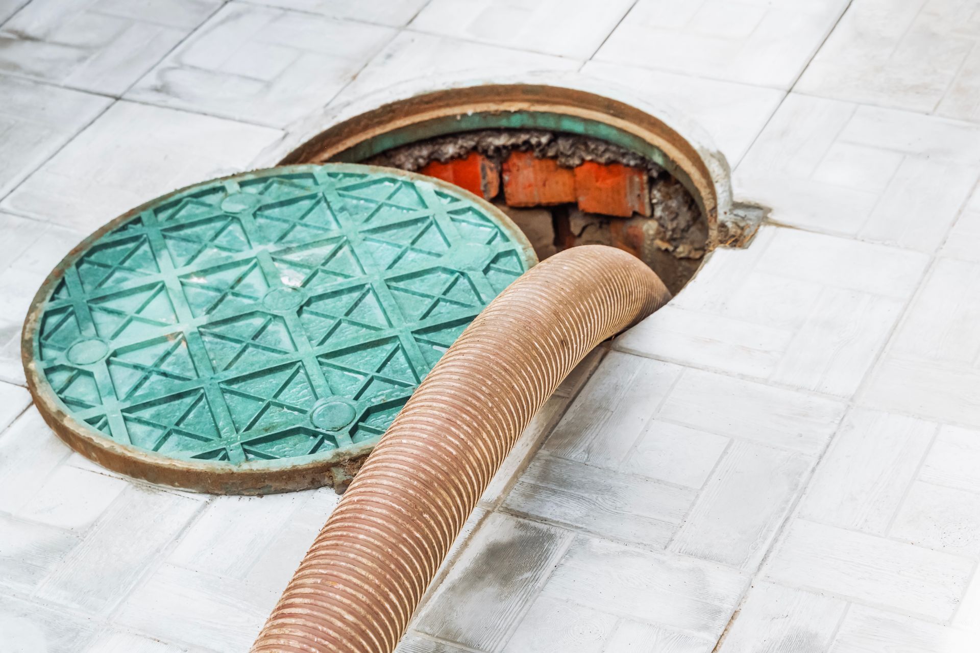 Open sewer access point with hose inside; green lid nearby on tiled floor. Brick interior.
