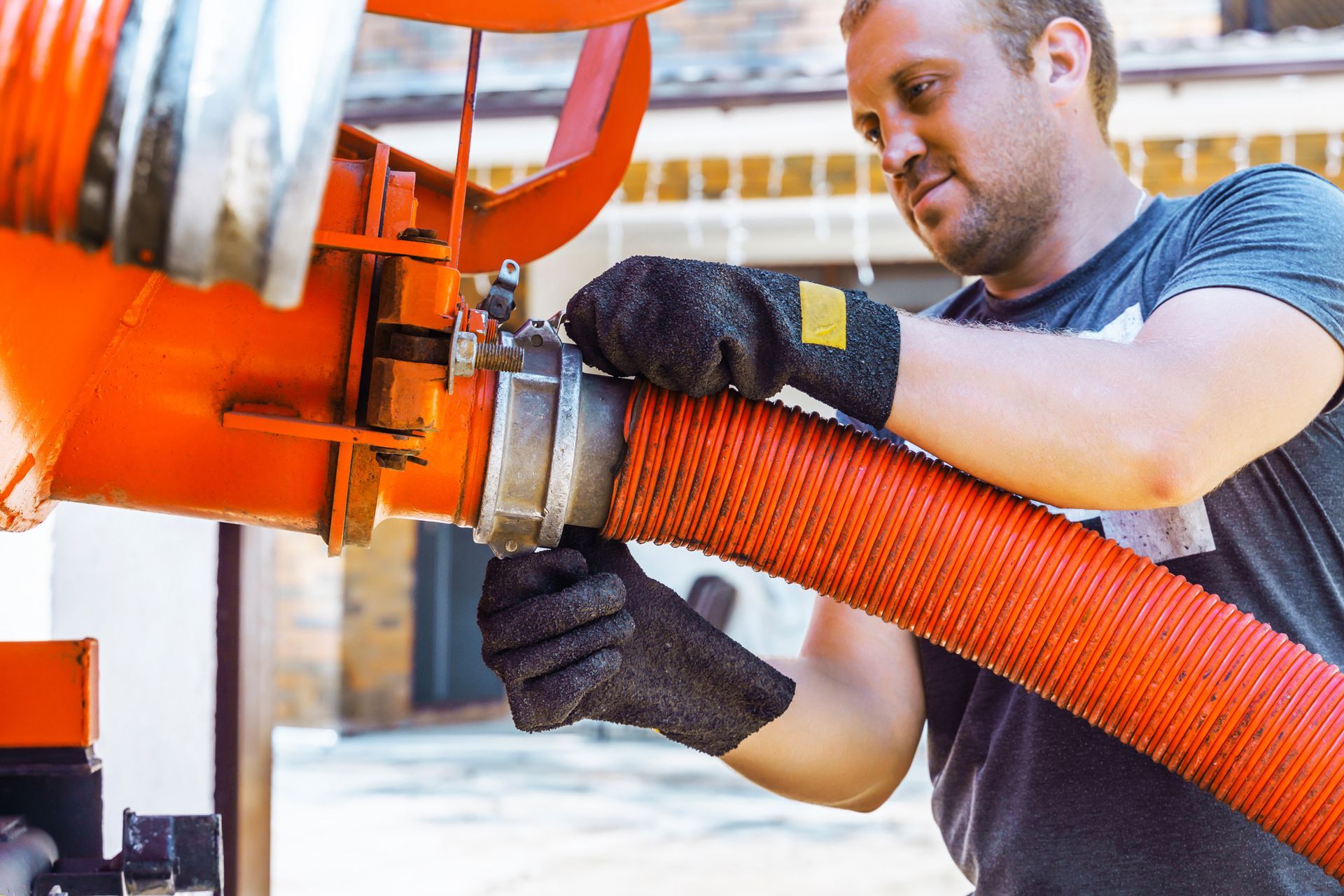 Man in gloves connects a large orange hose to machinery outdoors.