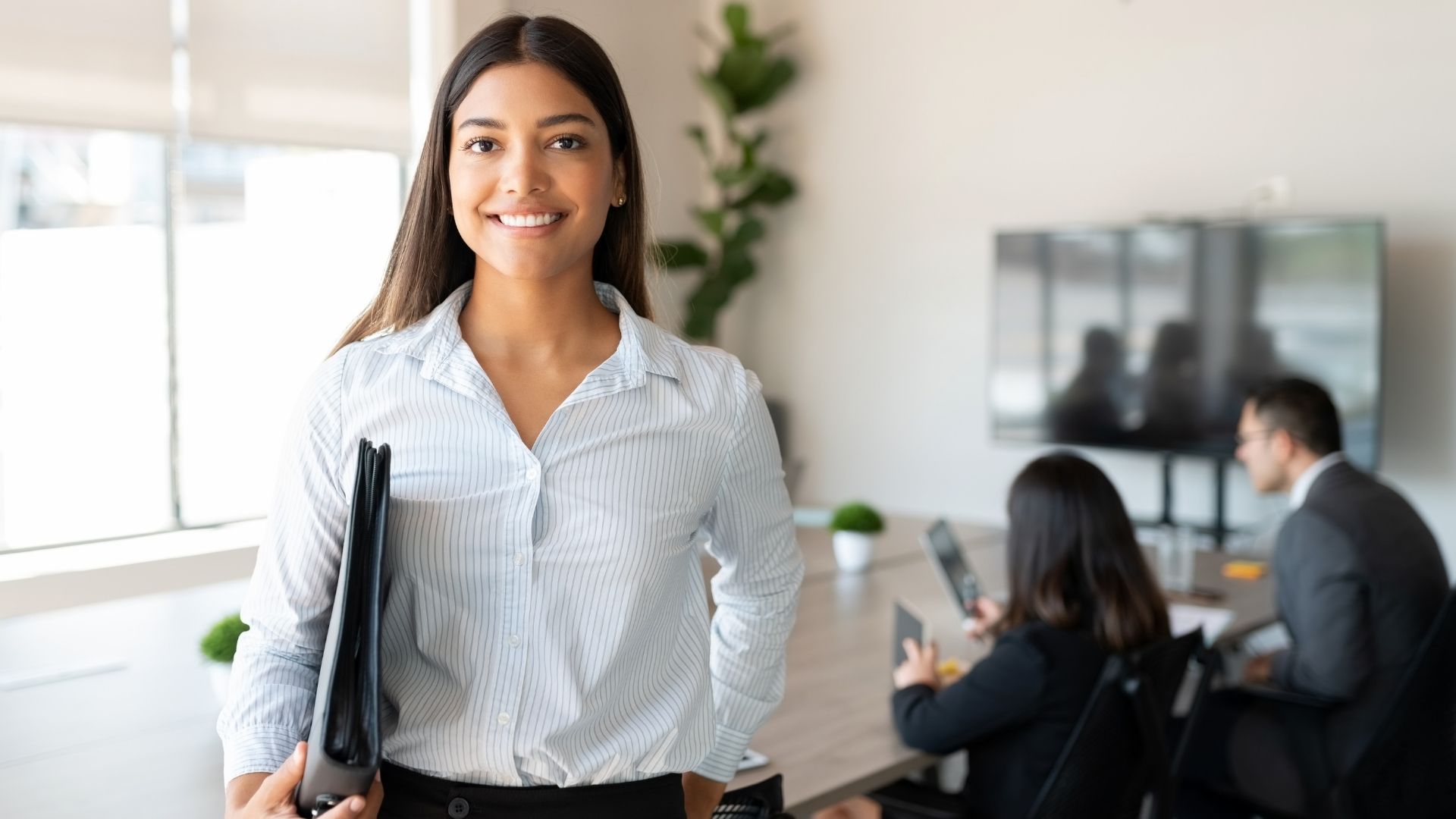 A woman is holding a folder and smiling in an office.