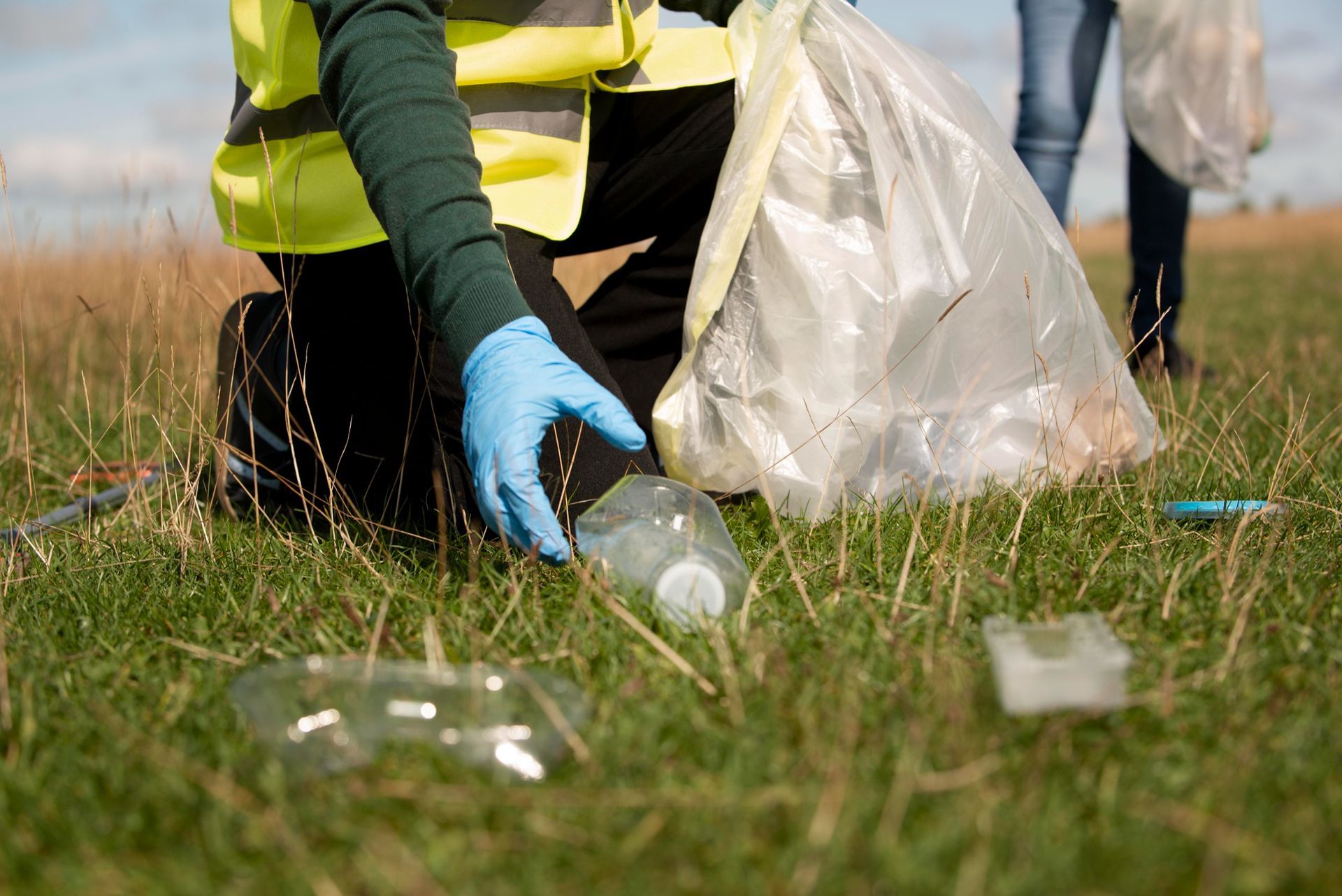 Persona recogiendo basura en un campo de hierba, vistiendo un chaleco de seguridad y guantes.