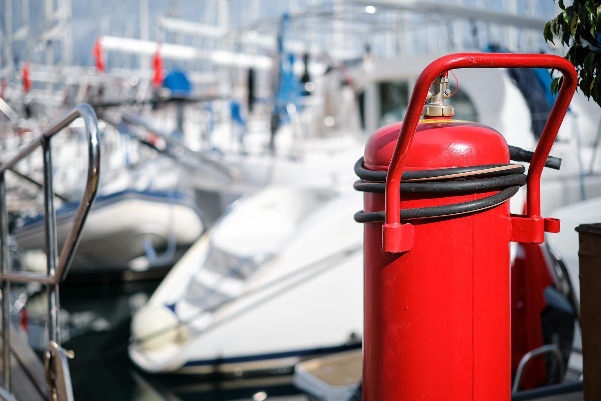 Tanque de propano rojo con manguera negra en un muelle de barcos, barcos borrosos en el fondo.
