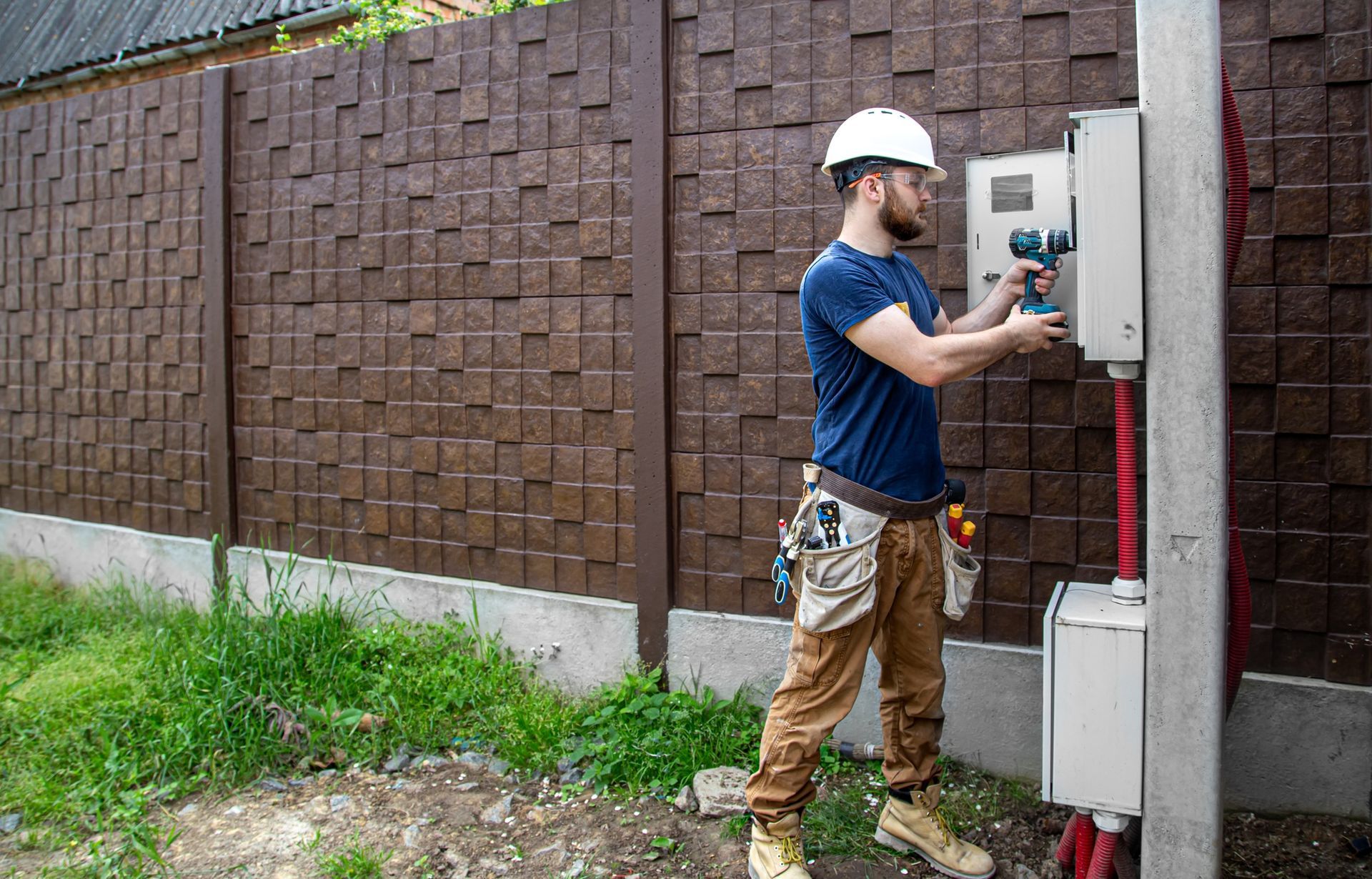 Un electricista con casco usa un taladro en una caja de servicios públicos, cerca de una cerca texturizada de color marrón.
