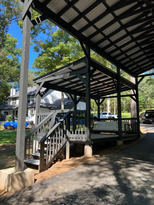 A black wooden outdoor pavilion with stairs and a seating area stands in a grassy backyard under trees.