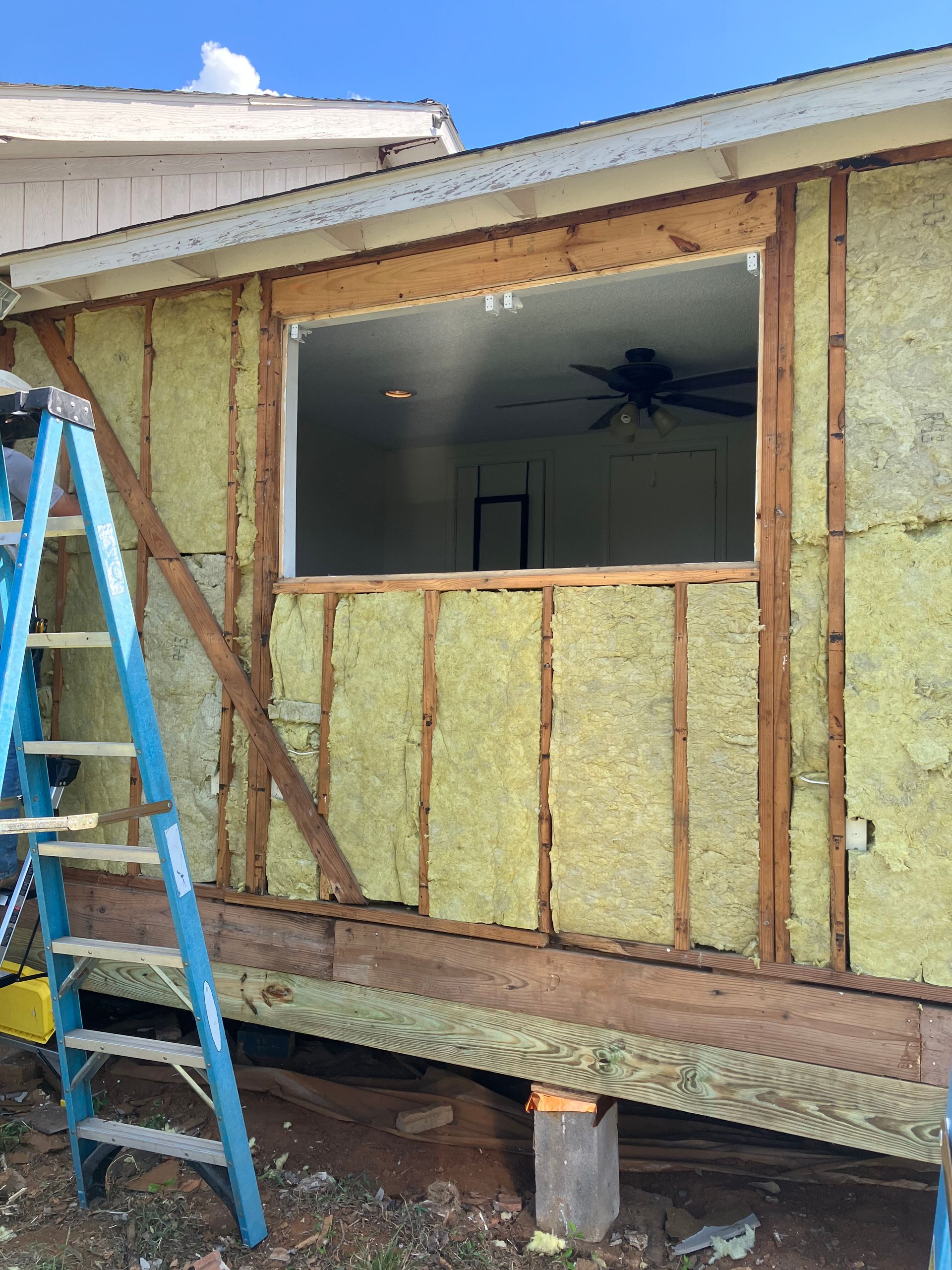 A blue ladder leans against the exterior wall of a house undergoing renovation, revealing yellow insulation and framing.