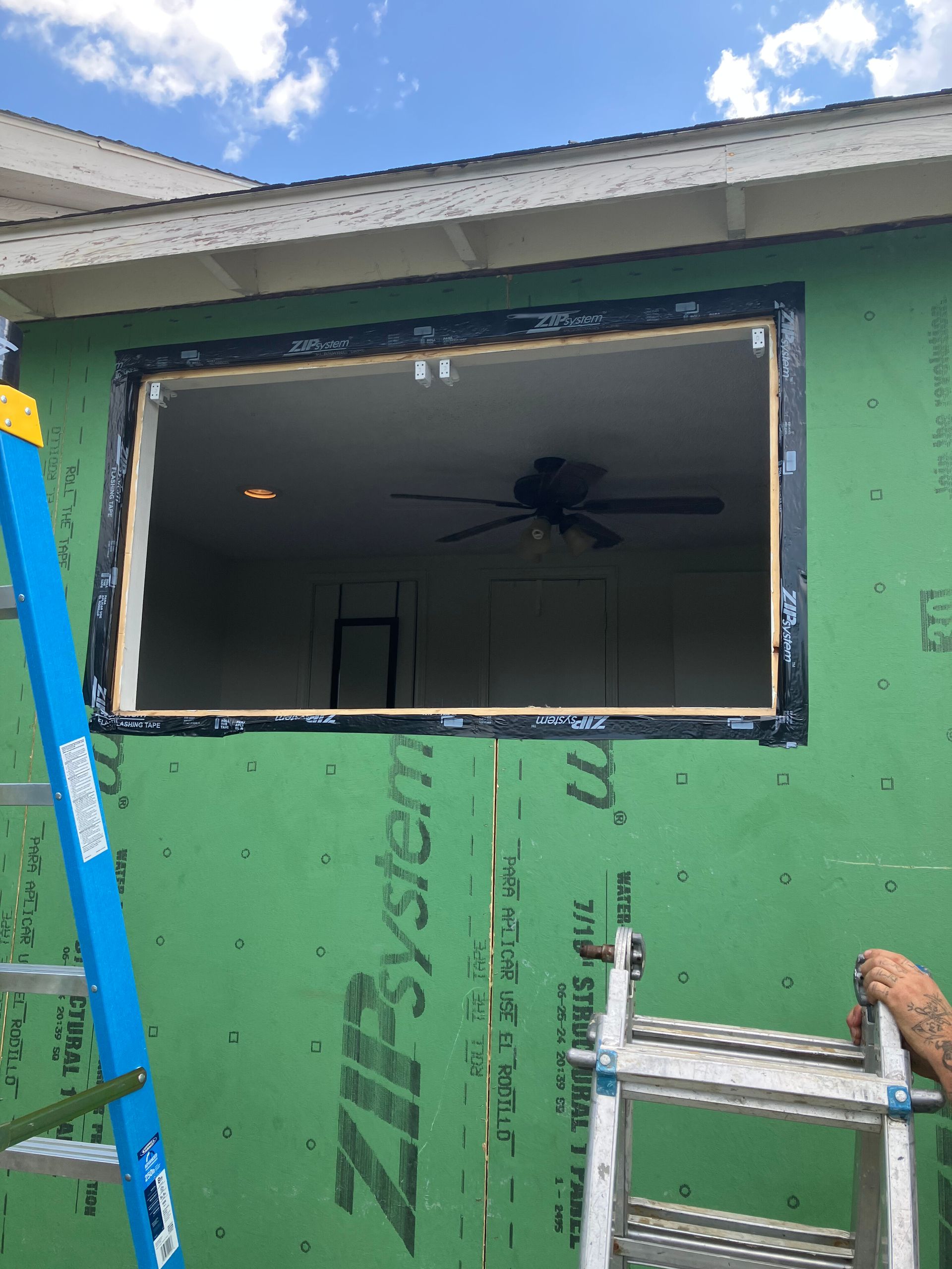 An open window frame installed in a wall covered with green Zip System sheathing, with a ladder visible in the foreground.