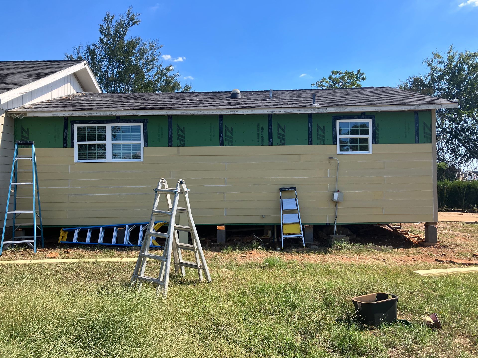 A side view of a house undergoing renovations with green sheathing, yellow siding, and multiple ladders against the wall.