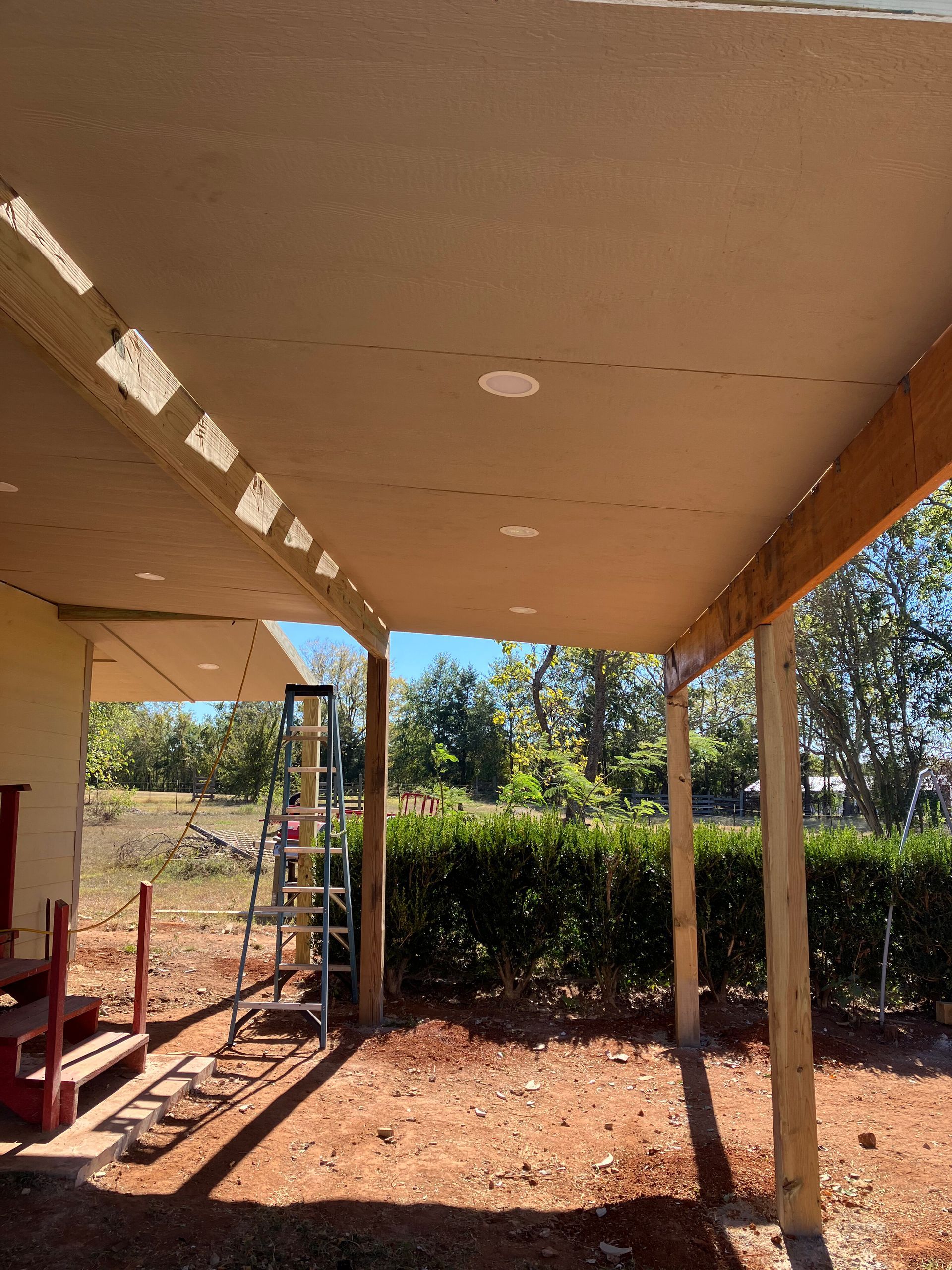 A tall ladder stands under a rustic, unfinished covered patio porch with wooden support posts in a yard.