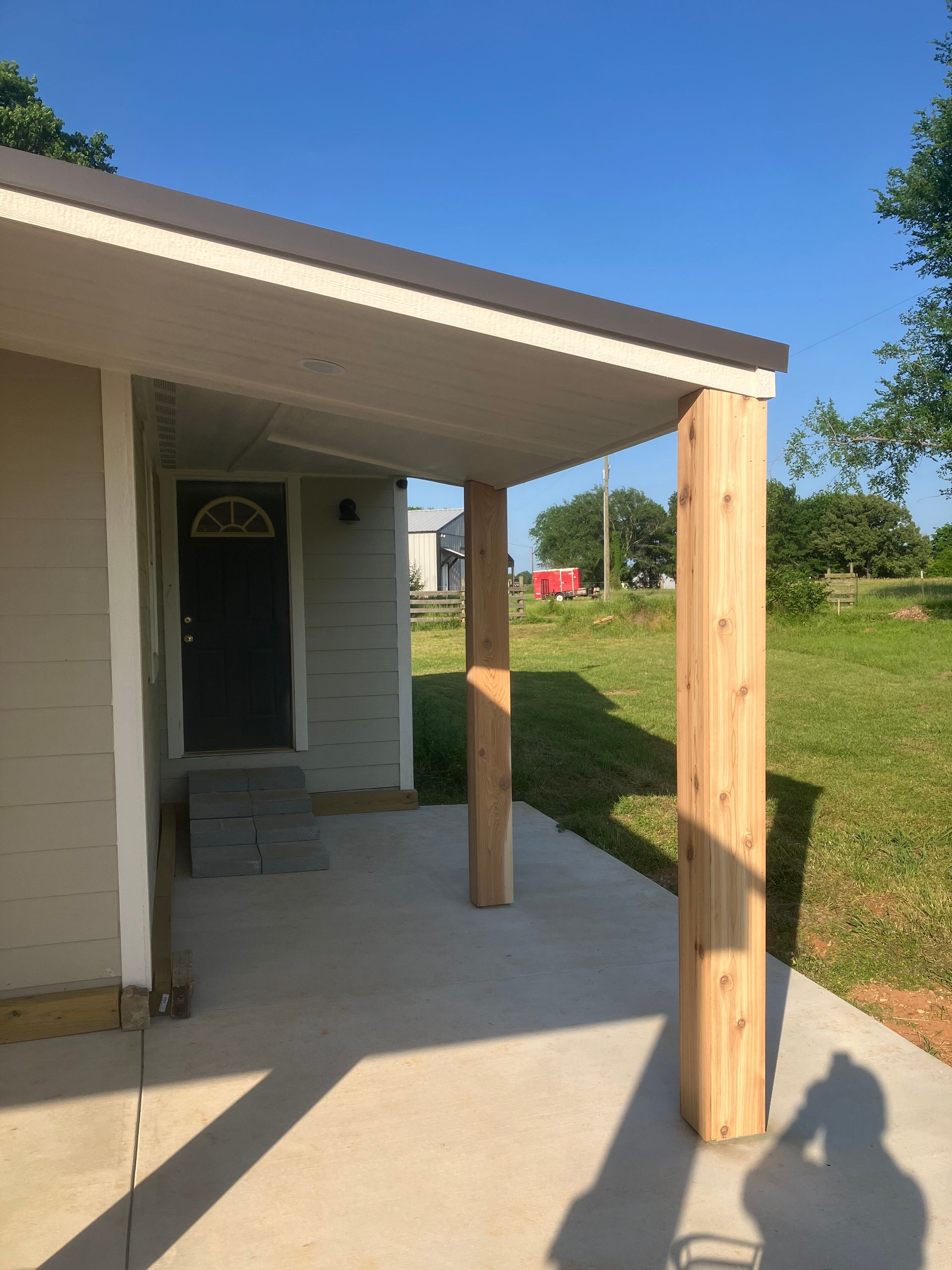 A beige house exterior with a concrete porch and two new vertical wooden support posts under a grey metal roof.