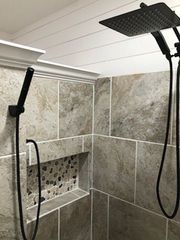 A modern walk-in shower with large beige stone tiles, a black rainfall shower head, a handheld sprayer, and a pebble-inlay.